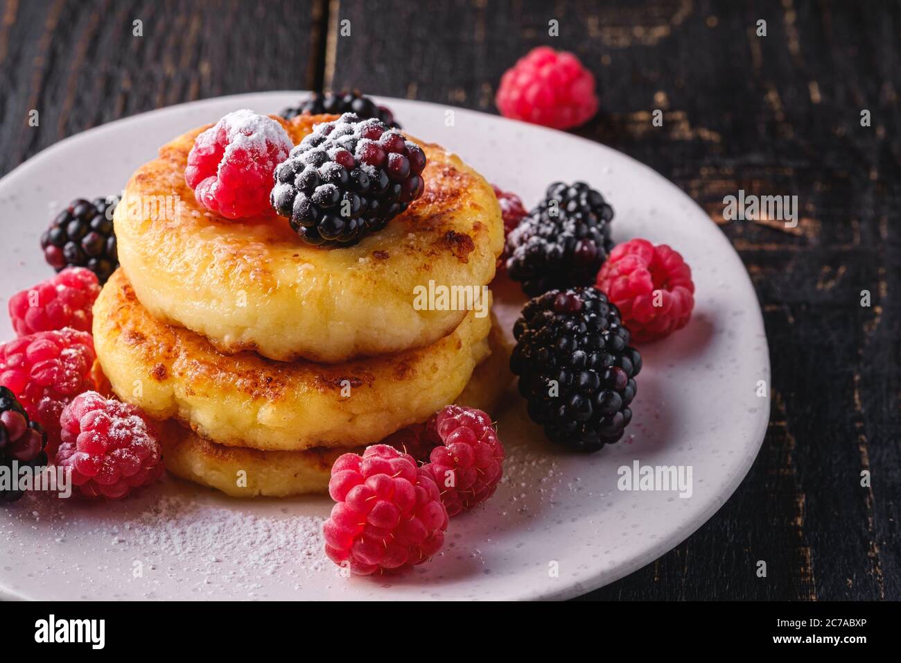 Frittelle di formaggio e zucchero in polvere, frittelle di cagliata dessert con lampone e frutti di bosco in piatto su sfondo nero scuro di legno Foto Stock