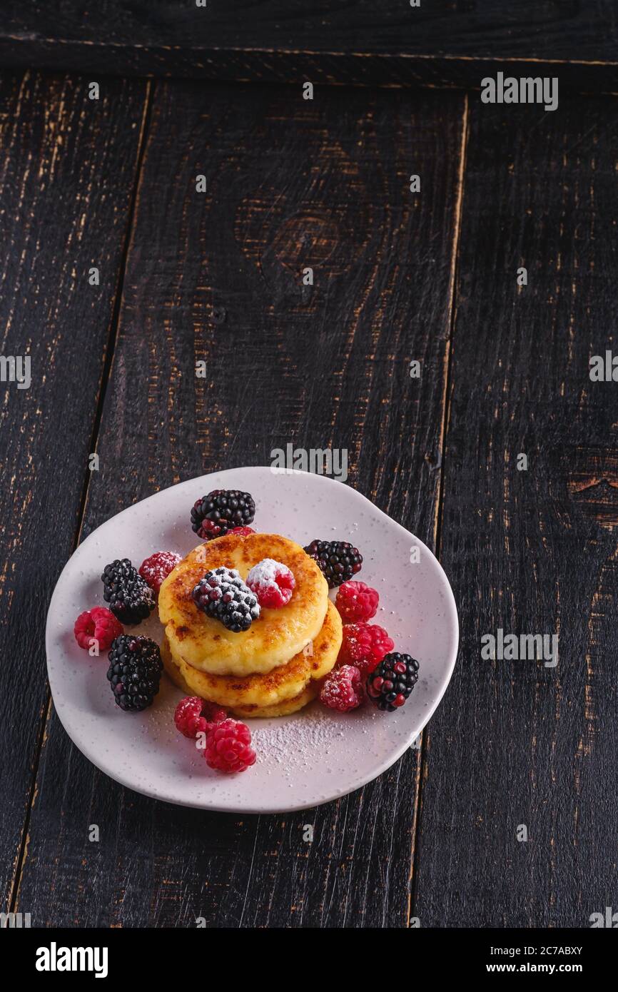 Frittelle di formaggio e zucchero in polvere, frittelle di cagliata dessert con lampone e frutti di bosco in piatto su sfondo nero scuro di legno Foto Stock