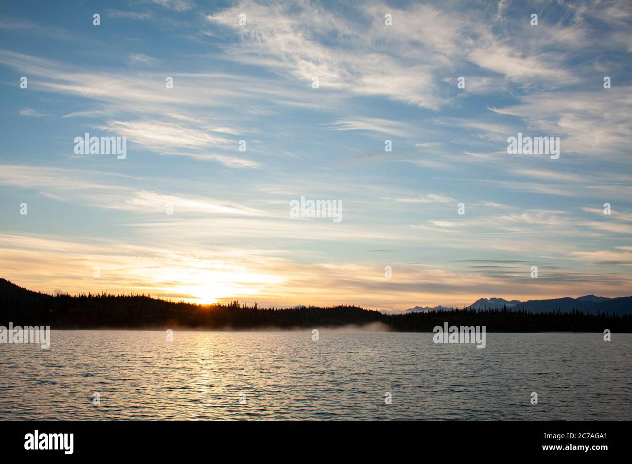 Tramonto dorato su un lago dell'Alaska, con sagome di montagna e morbide nuvole che riflettono la serena bellezza della natura selvaggia durante il crepuscolo. Foto Stock