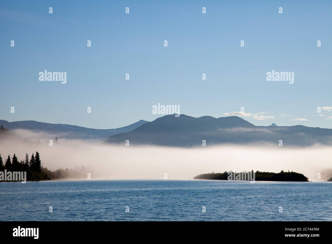 La nebbia mattutina si propaga attraverso il lago ICommanna, Alaska, con montagne sullo sfondo e luce del sole che si riflettono sulle acque tranquille e tranquille della natura selvaggia Foto Stock