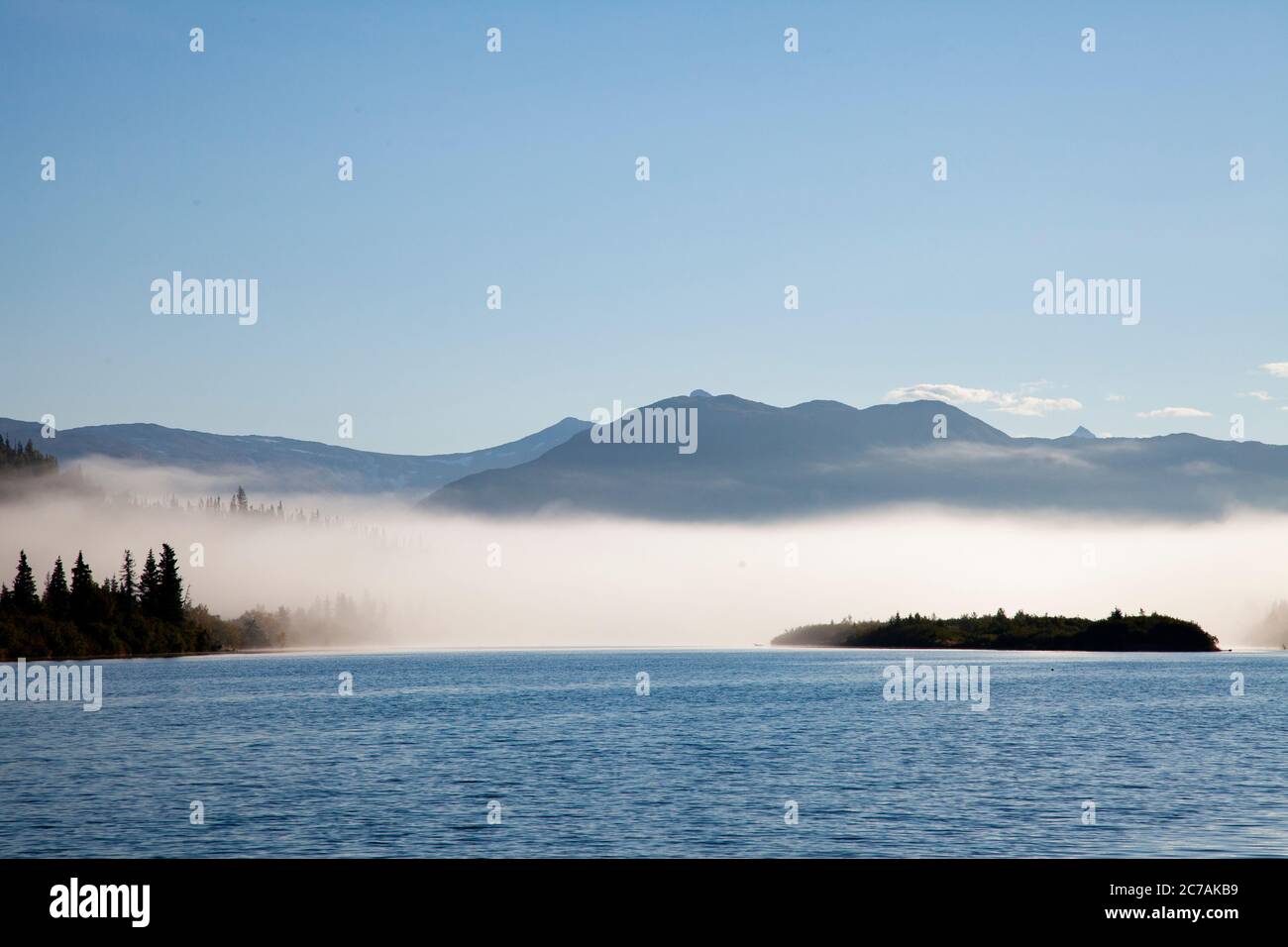 La nebbia mattutina si propaga attraverso il lago ICommanna, Alaska, con montagne sullo sfondo e luce del sole che si riflettono sulle acque tranquille e tranquille della natura selvaggia Foto Stock