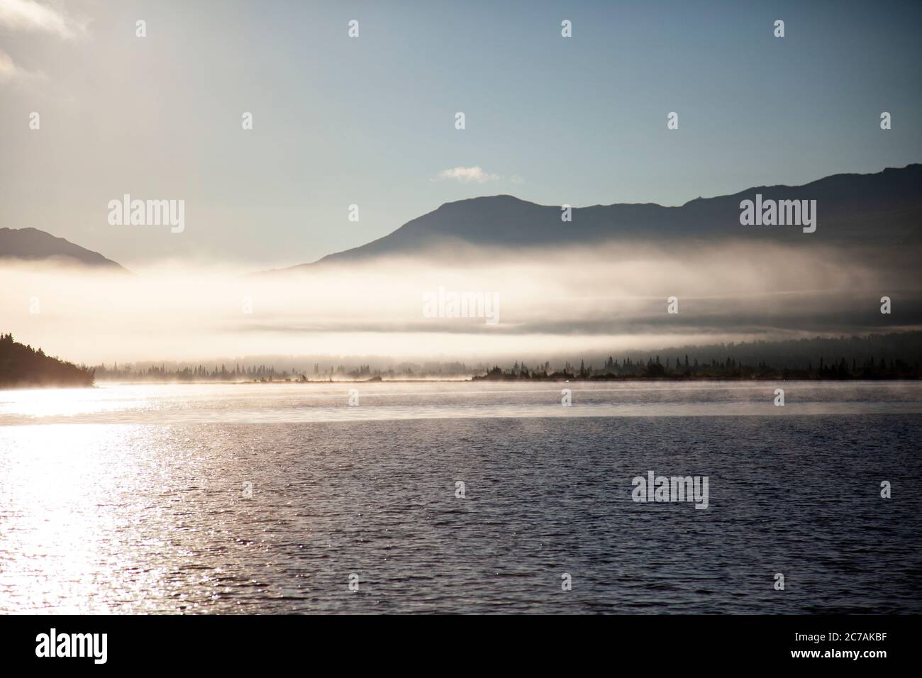 La nebbia mattutina si propaga attraverso il lago ICommanna, Alaska, con montagne sullo sfondo e luce del sole che si riflettono sulle acque tranquille e tranquille della natura selvaggia Foto Stock