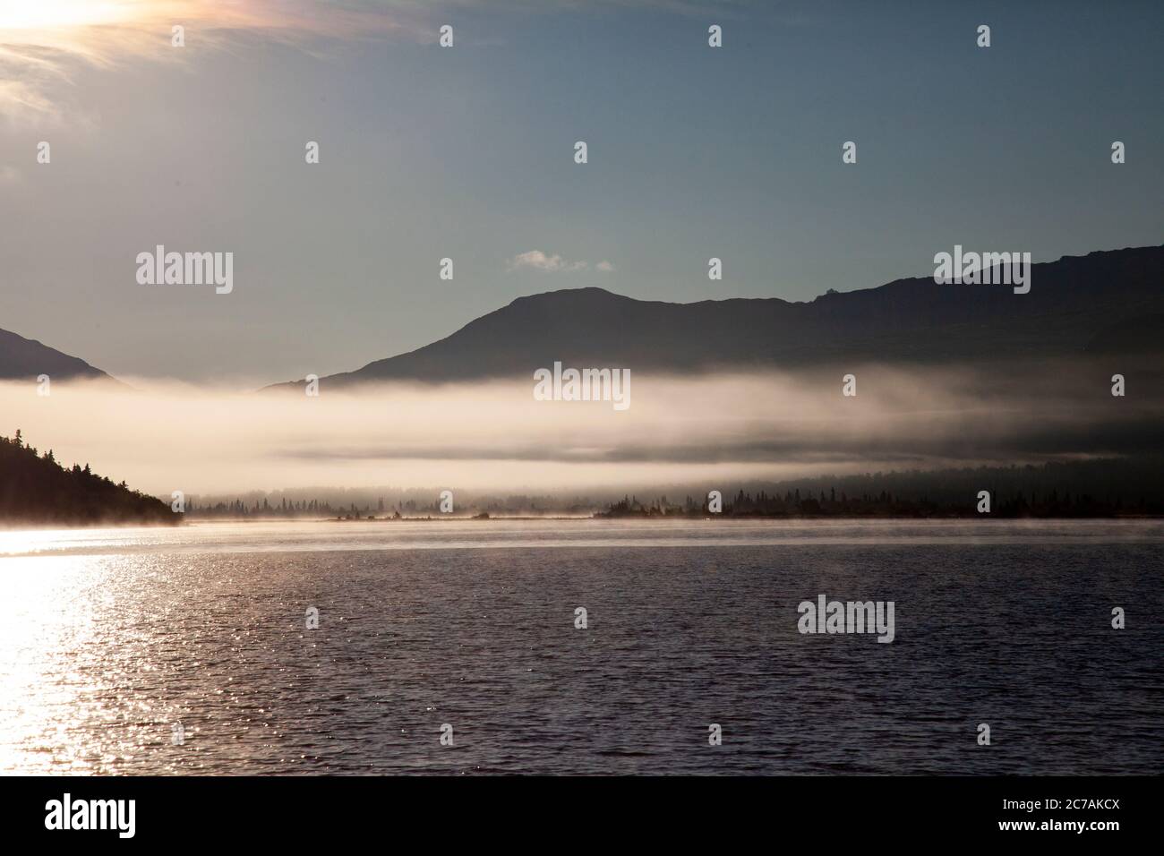 La nebbia mattutina si propaga attraverso il lago ICommanna, Alaska, con montagne sullo sfondo e luce del sole che si riflettono sulle acque tranquille e tranquille della natura selvaggia Foto Stock