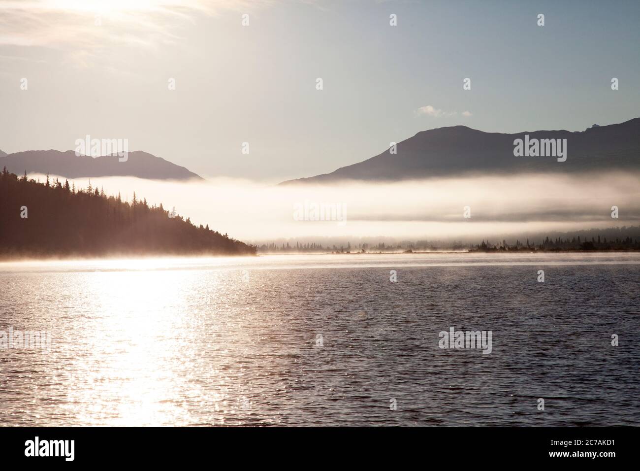 La nebbia mattutina si propaga attraverso il lago ICommanna, Alaska, con montagne sullo sfondo e luce del sole che si riflettono sulle acque tranquille e tranquille della natura selvaggia Foto Stock