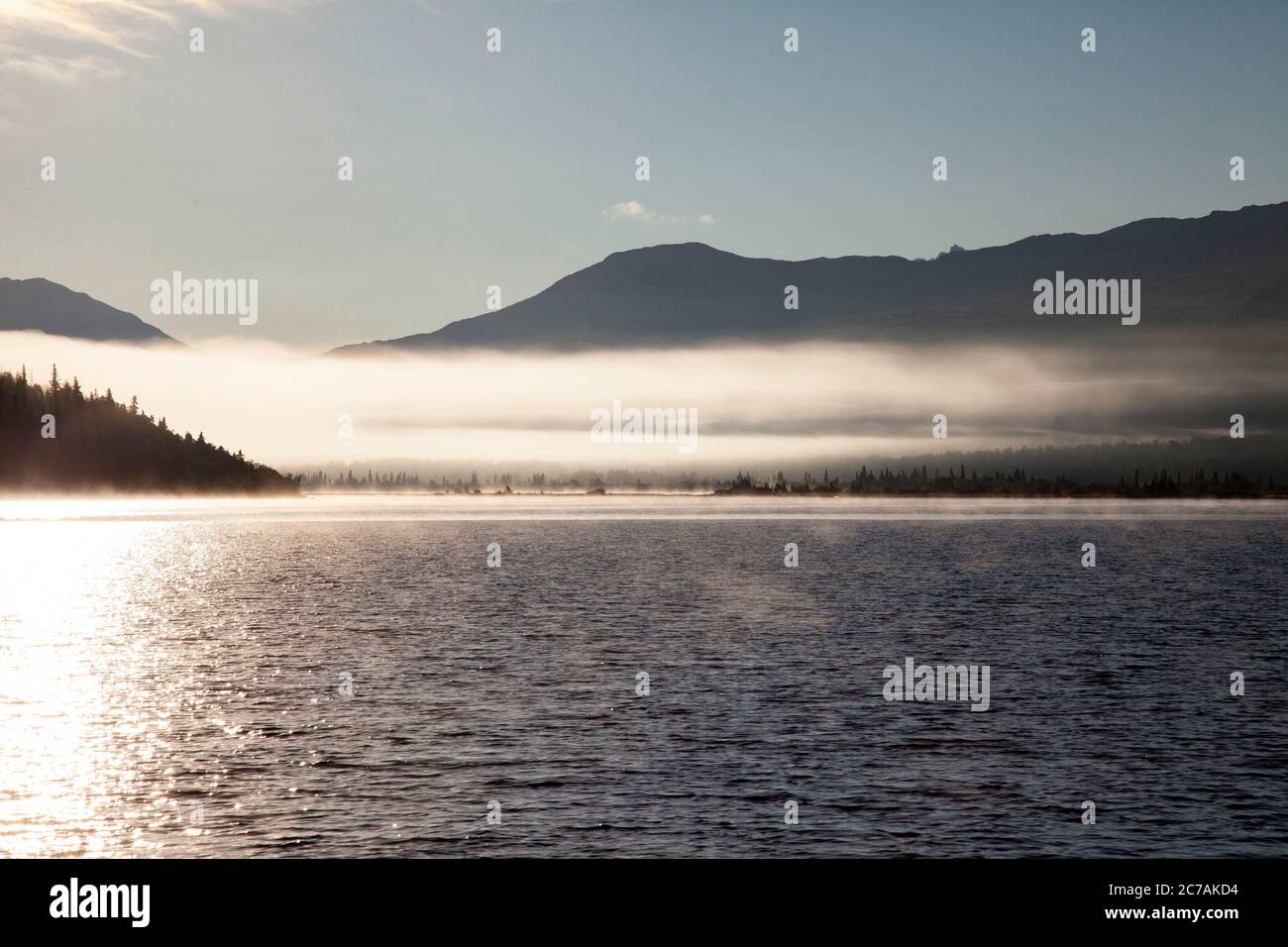 La nebbia mattutina si propaga attraverso il lago ICommanna, Alaska, con montagne sullo sfondo e luce del sole che si riflettono sulle acque tranquille e tranquille della natura selvaggia Foto Stock