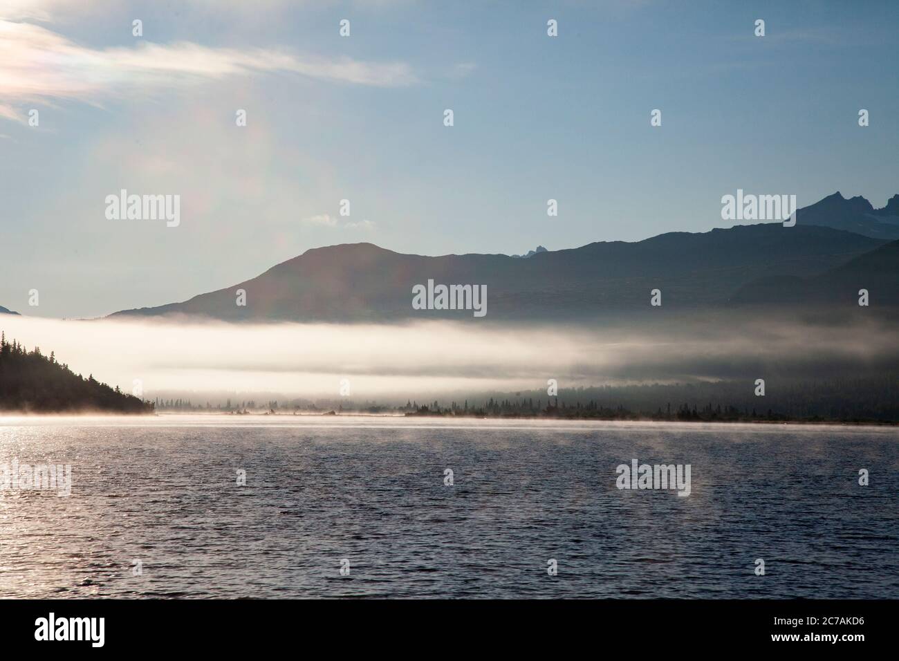 La nebbia mattutina si propaga attraverso il lago ICommanna, Alaska, con montagne sullo sfondo e luce del sole che si riflettono sulle acque tranquille e tranquille della natura selvaggia Foto Stock