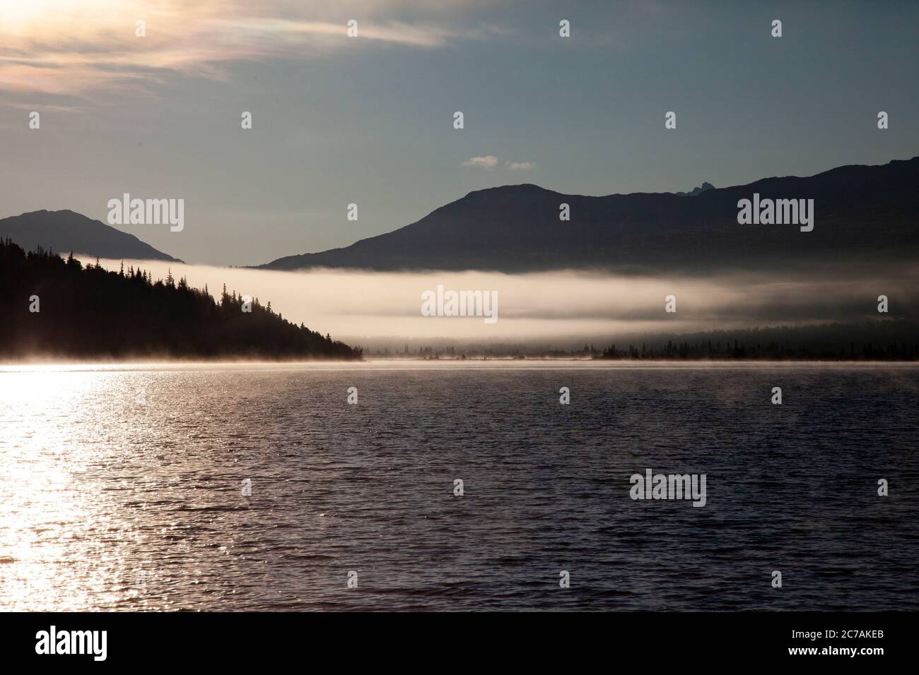 La nebbia mattutina si propaga attraverso il lago ICommanna, Alaska, con montagne sullo sfondo e luce del sole che si riflettono sulle acque tranquille e tranquille della natura selvaggia Foto Stock