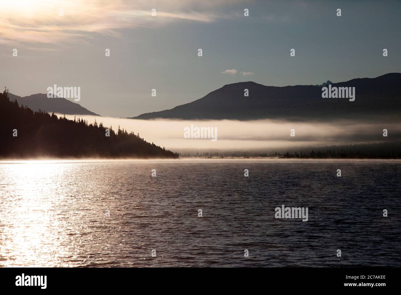 La nebbia mattutina si propaga attraverso il lago ICommanna, Alaska, con montagne sullo sfondo e luce del sole che si riflettono sulle acque tranquille e tranquille della natura selvaggia Foto Stock
