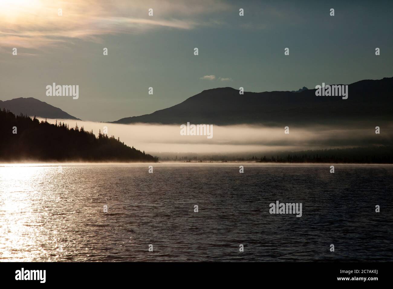La nebbia mattutina si propaga attraverso il lago ICommanna, Alaska, con montagne sullo sfondo e luce del sole che si riflettono sulle acque tranquille e tranquille della natura selvaggia Foto Stock