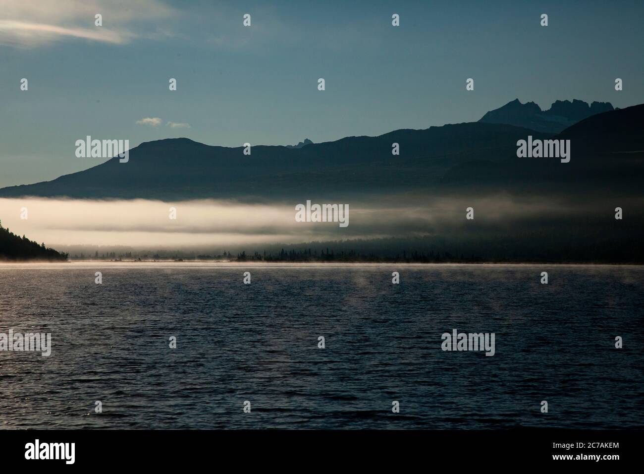 La nebbia mattutina si propaga attraverso il lago ICommanna, Alaska, con montagne sullo sfondo e luce del sole che si riflettono sulle acque tranquille e tranquille della natura selvaggia Foto Stock