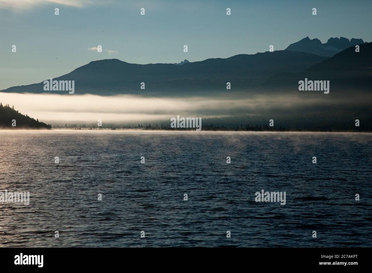 La nebbia mattutina si propaga attraverso il lago ICommanna, Alaska, con montagne sullo sfondo e luce del sole che si riflettono sulle acque tranquille e tranquille della natura selvaggia Foto Stock