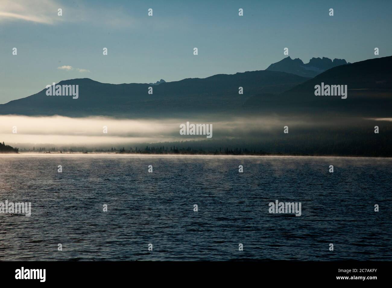 La nebbia mattutina si propaga attraverso il lago ICommanna, Alaska, con montagne sullo sfondo e luce del sole che si riflettono sulle acque tranquille e tranquille della natura selvaggia Foto Stock