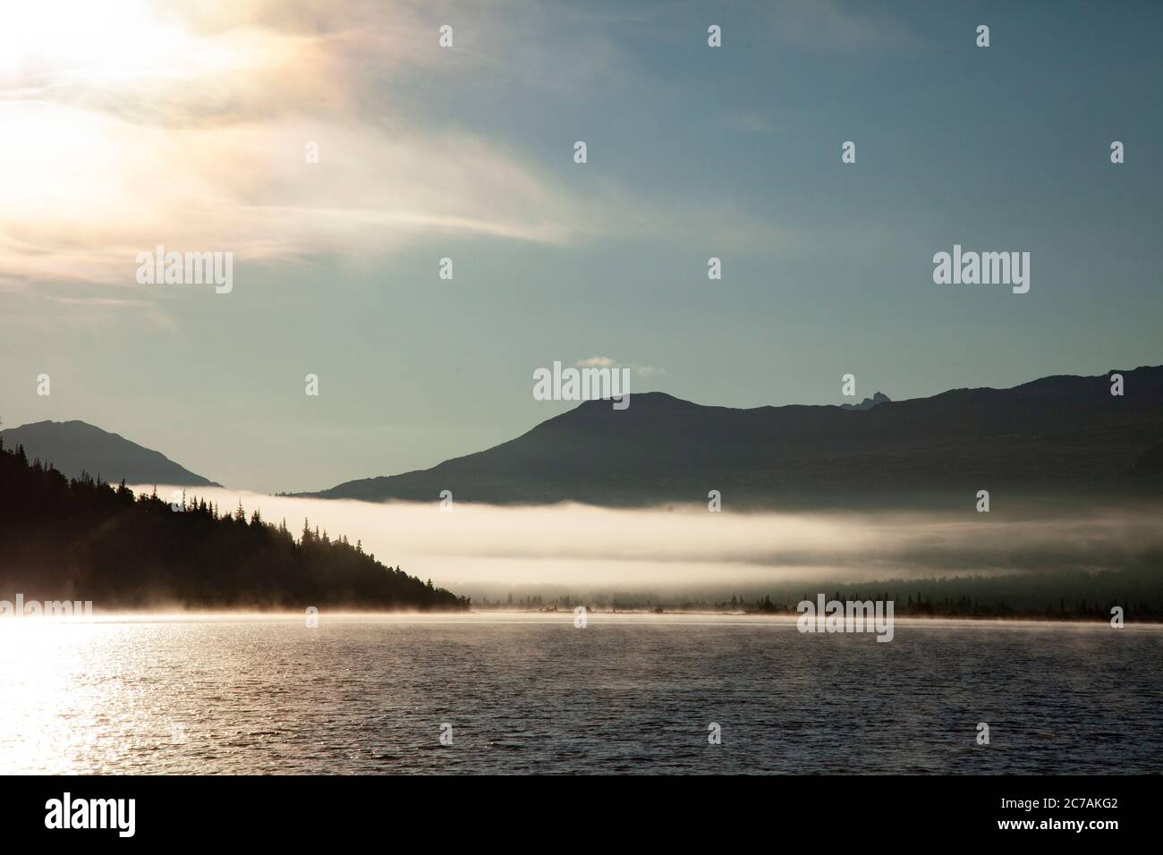 La nebbia mattutina si propaga attraverso il lago ICommanna, Alaska, con montagne sullo sfondo e luce del sole che si riflettono sulle acque tranquille e tranquille della natura selvaggia Foto Stock