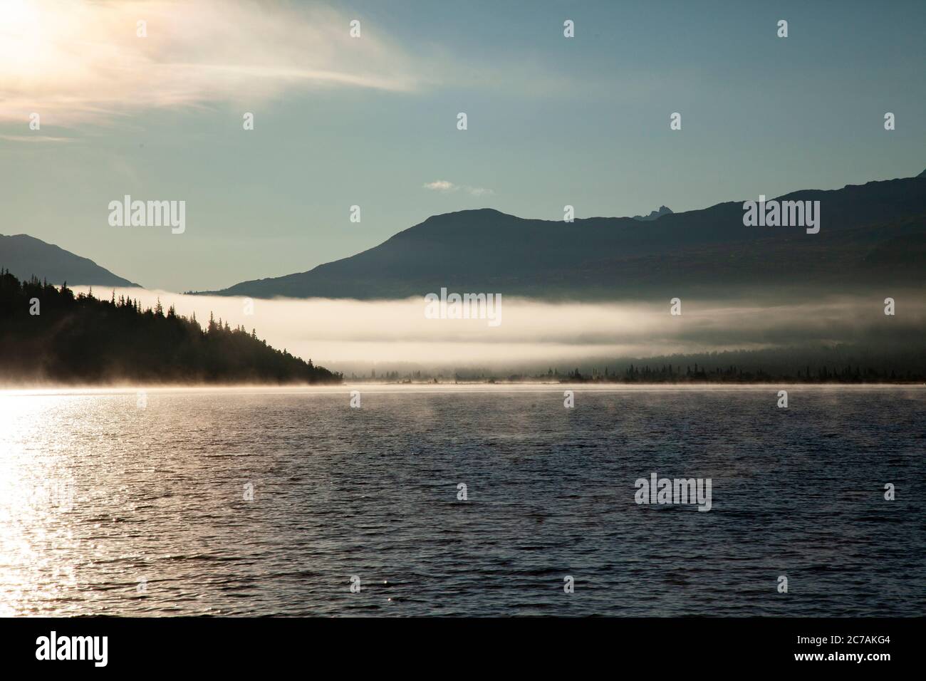 La nebbia mattutina si propaga attraverso il lago ICommanna, Alaska, con montagne sullo sfondo e luce del sole che si riflettono sulle acque tranquille e tranquille della natura selvaggia Foto Stock