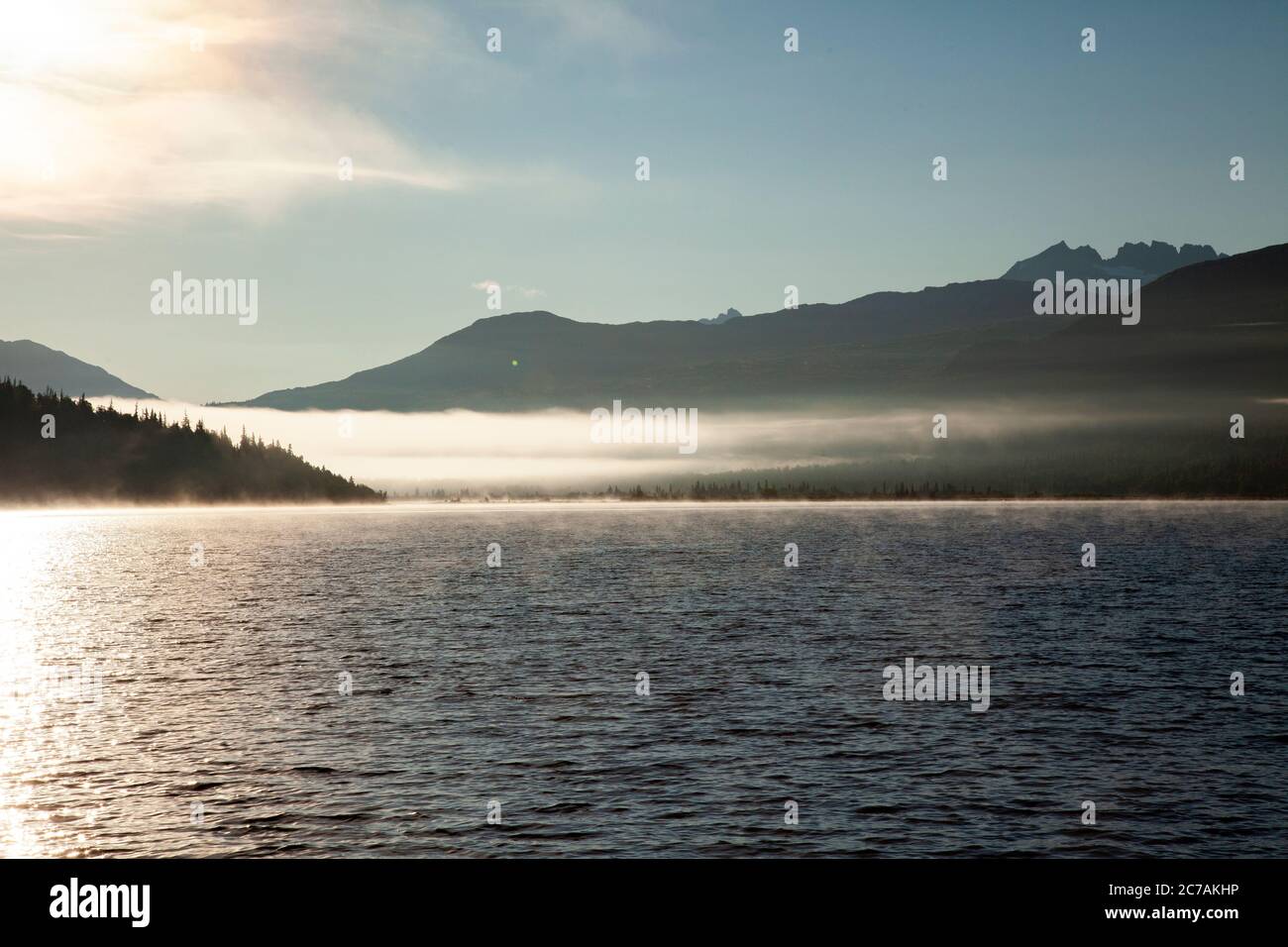 La nebbia mattutina si propaga attraverso il lago ICommanna, Alaska, con montagne sullo sfondo e luce del sole che si riflettono sulle acque tranquille e tranquille della natura selvaggia Foto Stock
