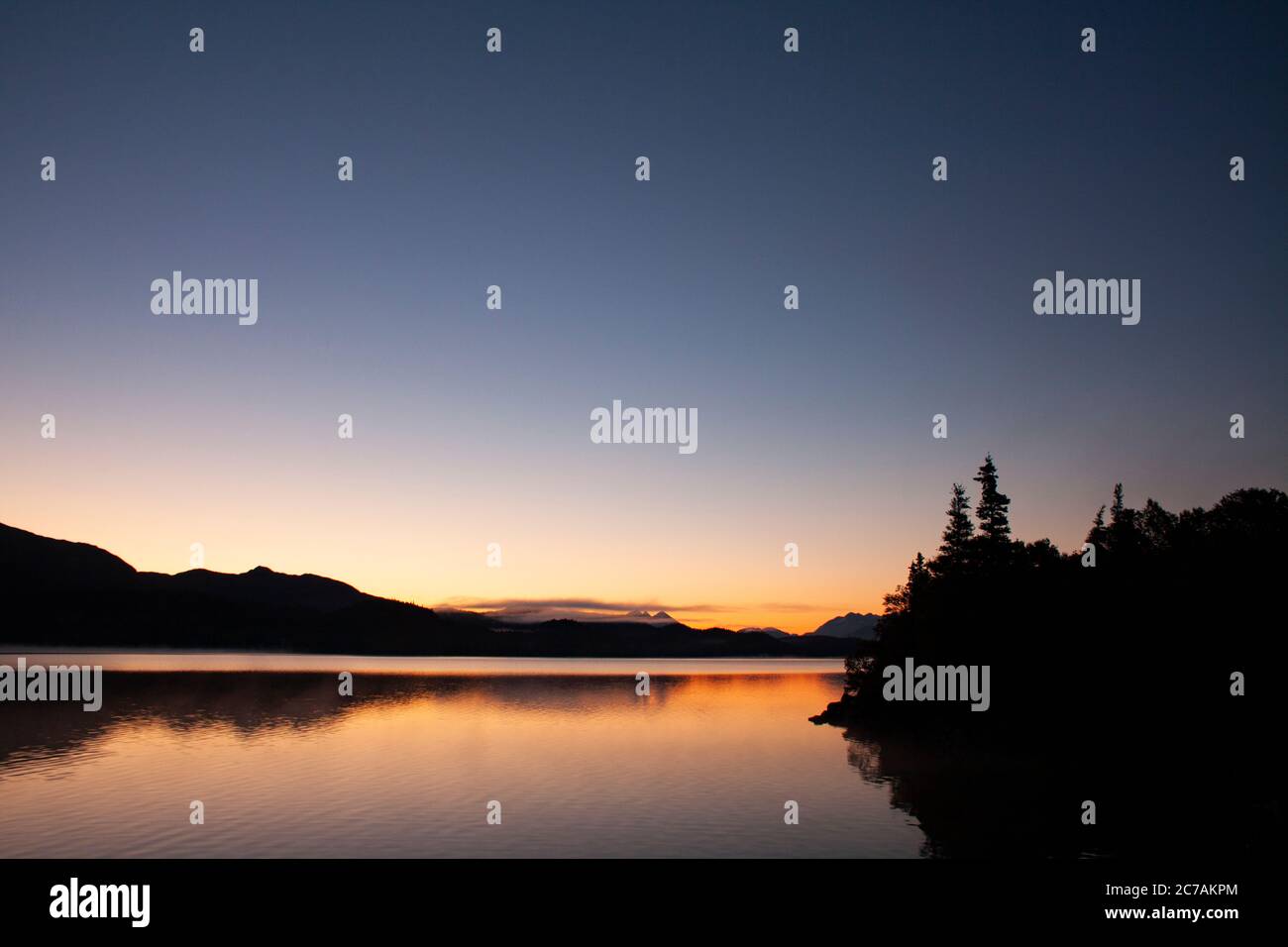 Alba tranquilla sul lago ICommanna, Alaska, con acque calme che riflettono il bagliore tenue dell'alba e le lontane sagome delle montagne nella natura selvaggia. Foto Stock