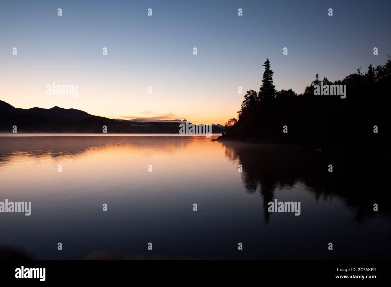 Alba tranquilla sul lago ICommanna, Alaska, con acque calme che riflettono il bagliore tenue dell'alba e le lontane sagome delle montagne nella natura selvaggia. Foto Stock