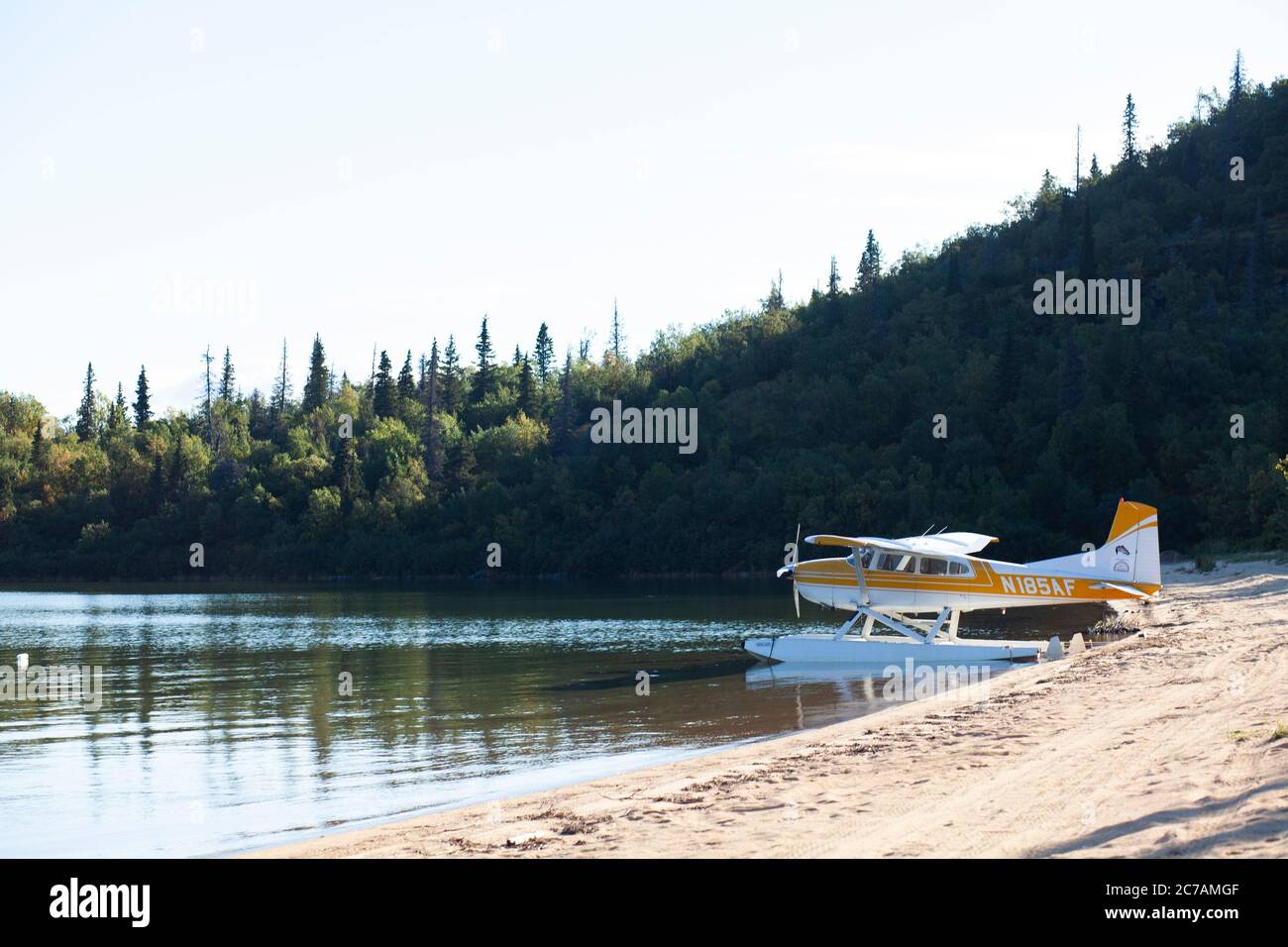 Idrovolante giallo attraccato sulle rive sabbiose del lago Iliamna, Alaska, circondato da lussureggianti colline boscose che riflettono cieli blu incontaminati. Foto Stock