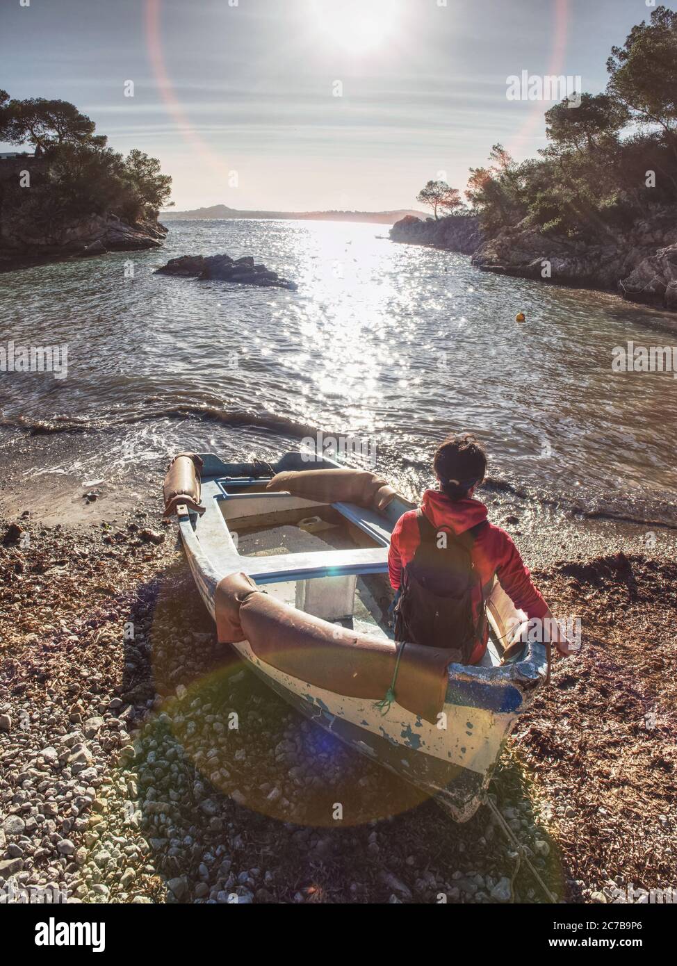 Giovane donna siede in barca sulla riva del mare e sogna. Baia di mare sassosa, vecchia barca da pesca è ancorata sulla ghiaia. Ragazza escursionista con zaino. Foto Stock
