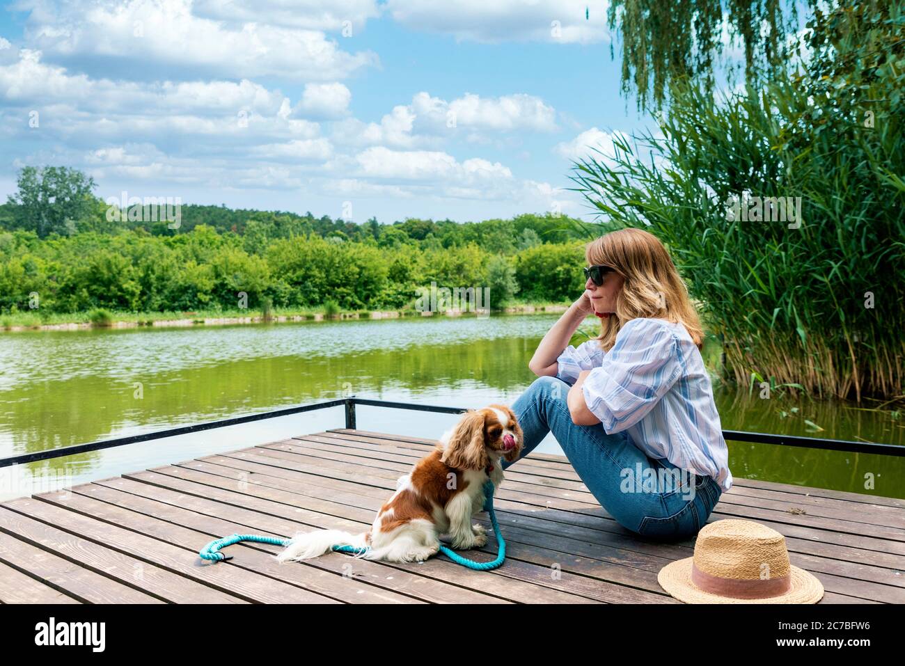 Foto a lunghezza intera di donna che sogna il giorno seduto sul molo accanto al suo cucciolo mentre si rilassa al lago. Foto Stock