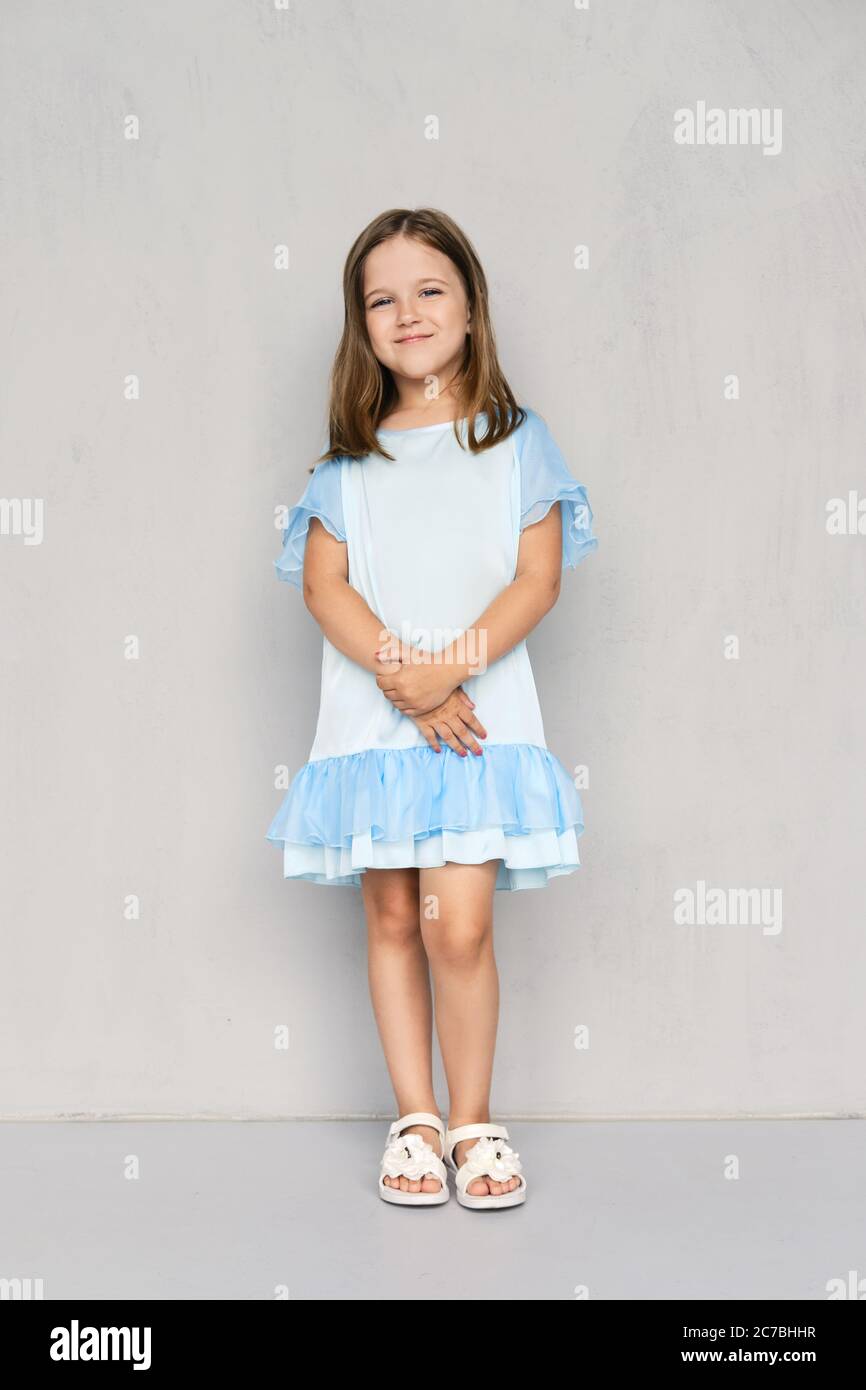 Carina bambina in abito blu e sandali bianchi in posa con le mani aggrappate vicino alla parete grigia Foto Stock