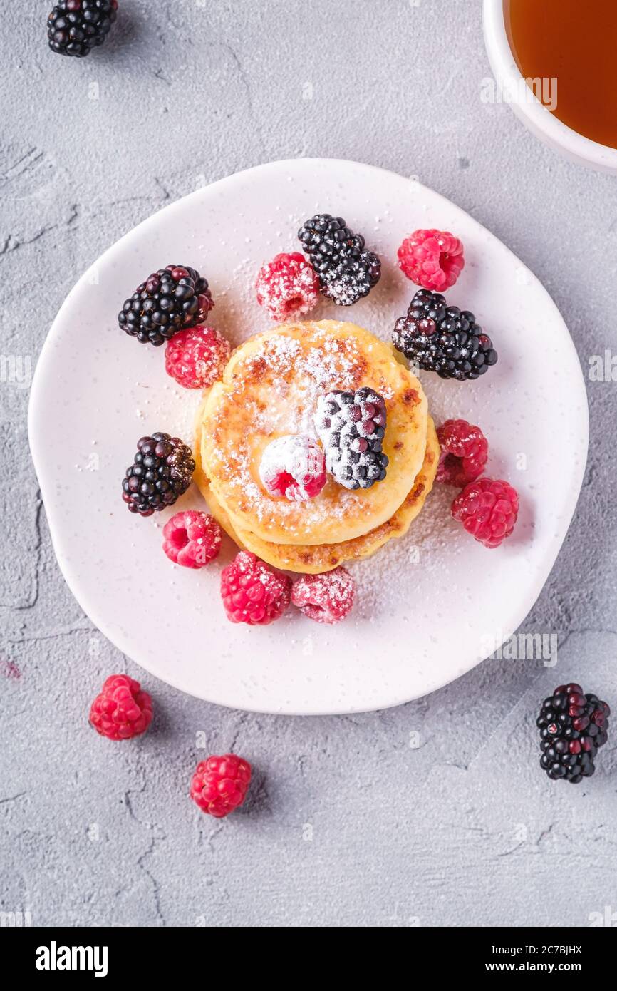 Frittelle al formaggio e zucchero in polvere, frittelle di cagliata dessert con lampone e frutti di bosco in piatto vicino alla tazza di tè caldo con fetta di limone Foto Stock