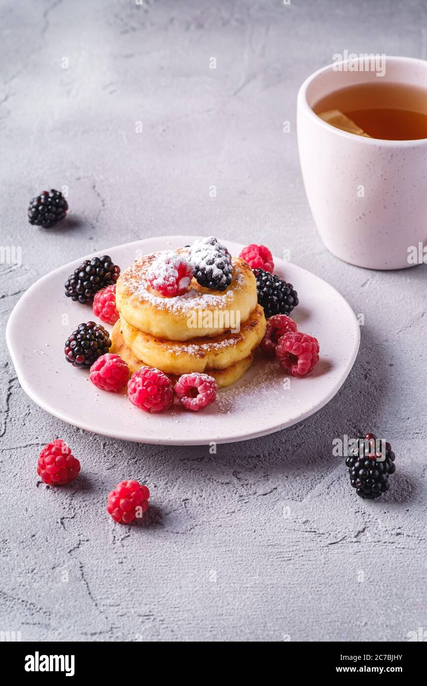 Frittelle al formaggio e zucchero in polvere, frittelle di cagliata dessert con lampone e frutti di bosco in piatto vicino alla tazza di tè caldo con fetta di limone Foto Stock