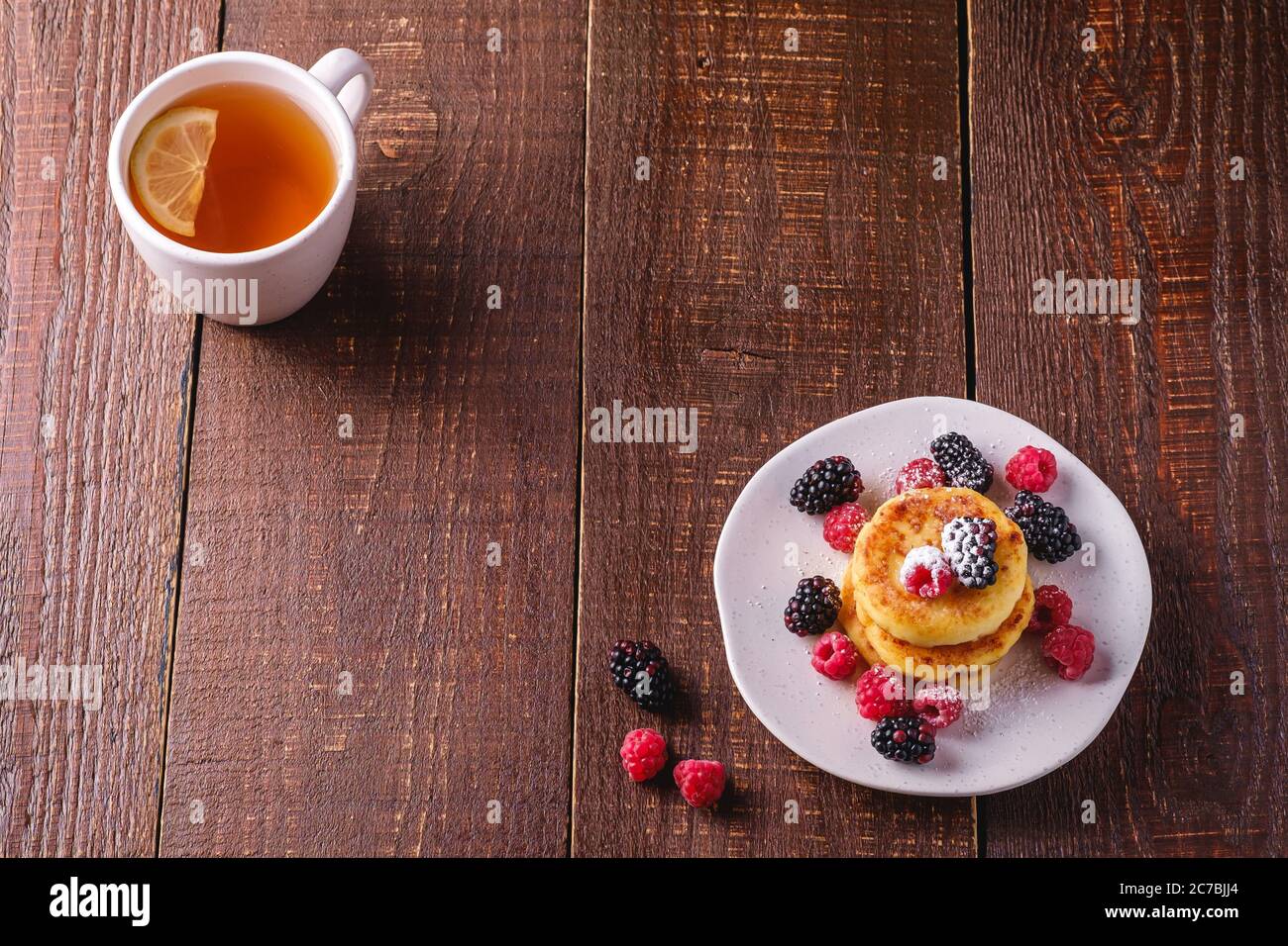 Frittelle di formaggio cottage, frittelle di cagliata dessert con lampone e frutti di bosco in piatto vicino alla tazza di tè caldo con fetta di limone Foto Stock