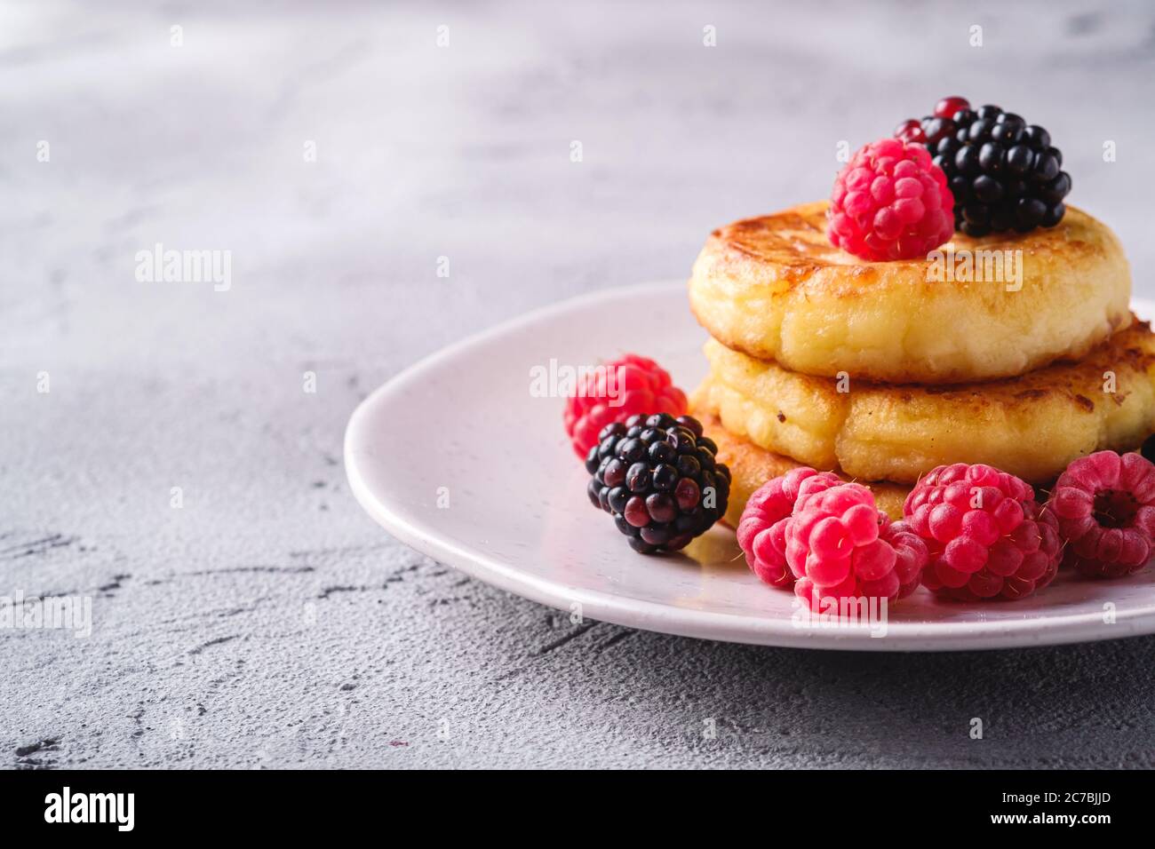 Frittelle al formaggio di cottage, frittelle di cagliata dessert con lampone e frutti di mora in piastra su fondo di pietra in cemento, macro angolo vista Foto Stock