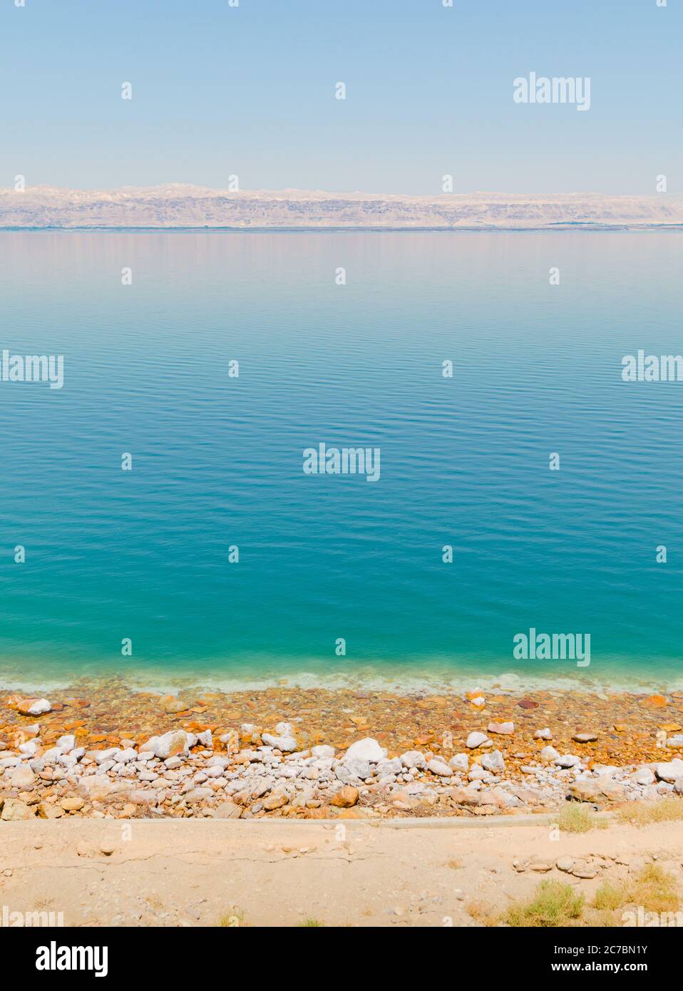Vista panoramica verticale della riva del Mar Morto, con crosta di sale e spiaggia di ghiaia vicino a un resort turistico, e la linea di montagna dei territori d'Israele sulla riva Foto Stock