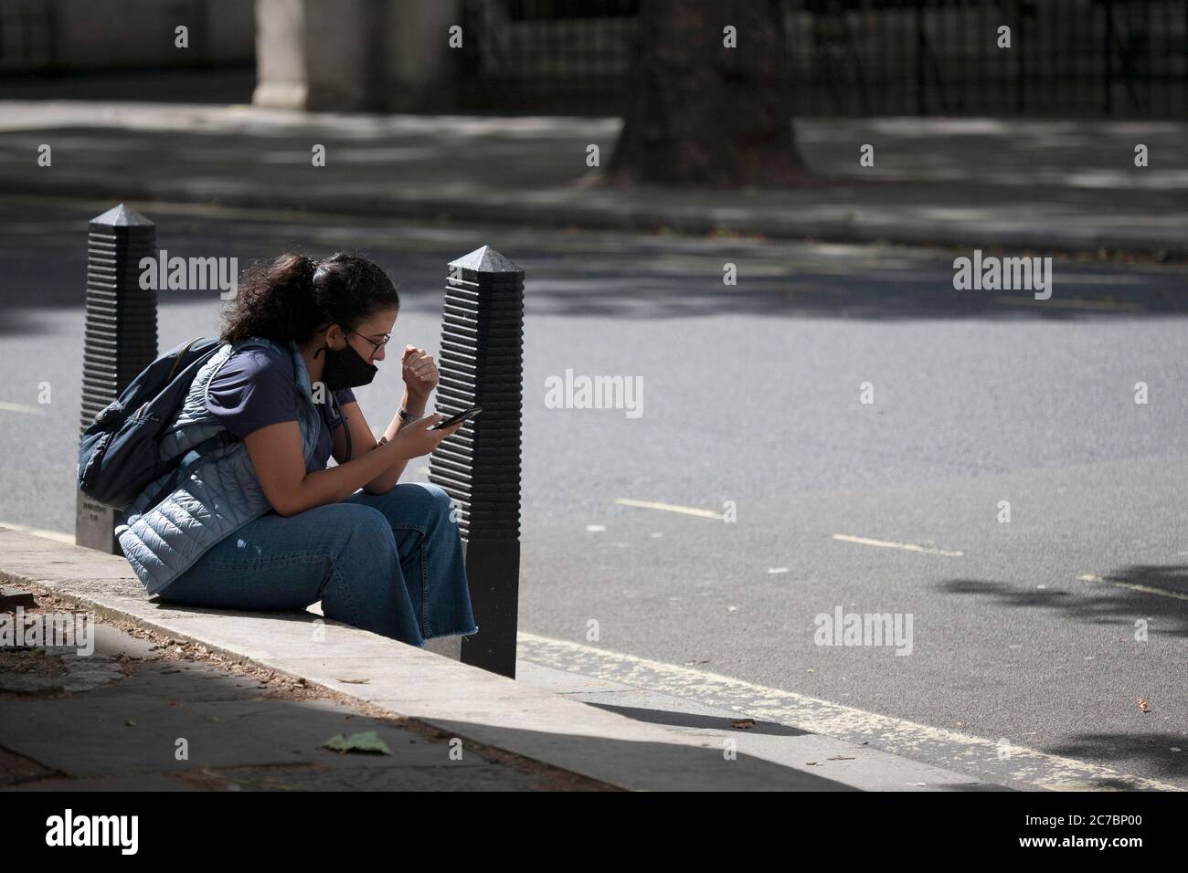 Giovane donna con un ponytail che indossa una maschera di faccia siede a bordo strada guardando il suo telefono fuori dei tribunali reali di giustizia. Londra, Inghilterra, Regno Unito Foto Stock