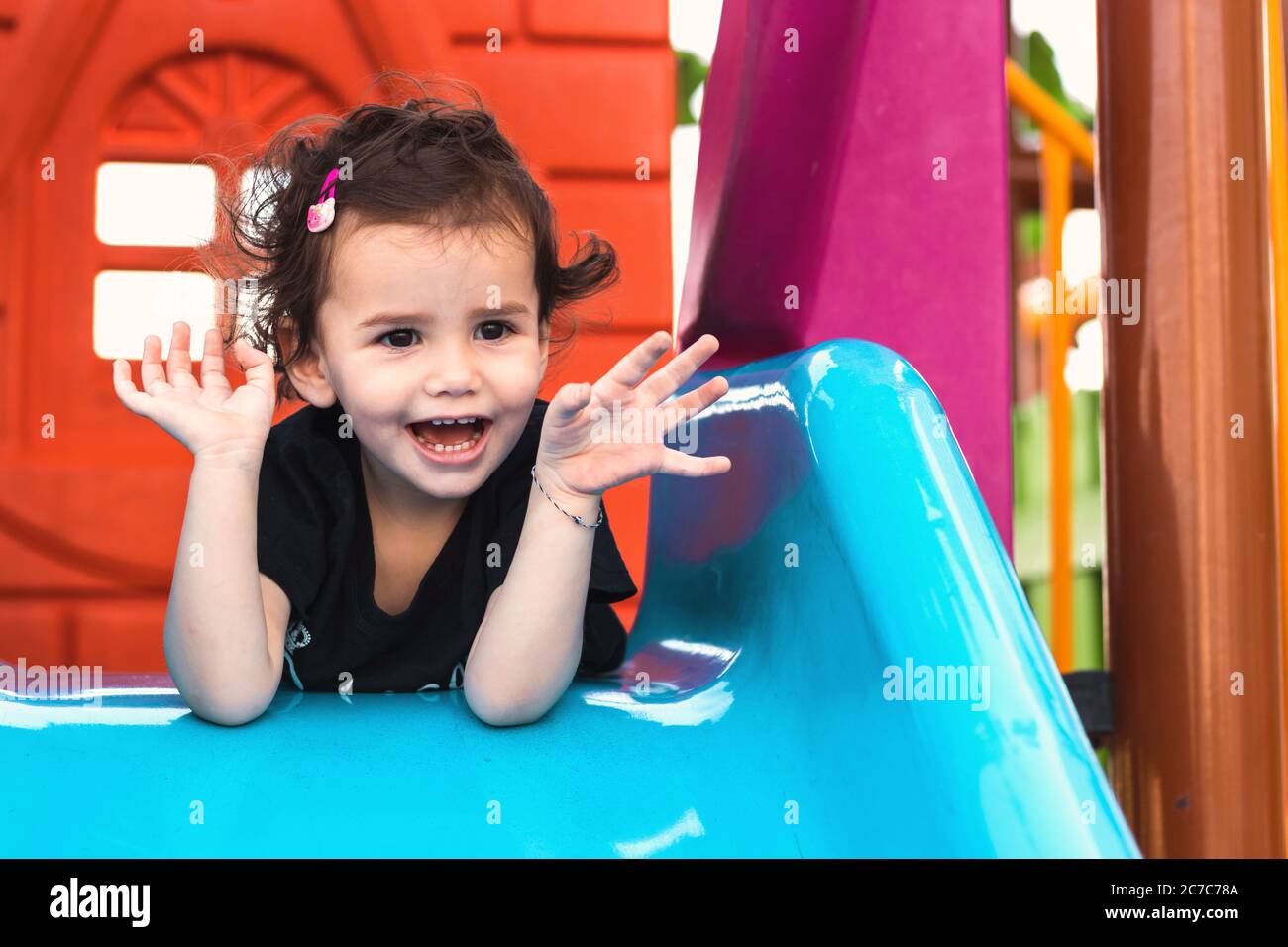 una ragazza liitle carina che si posa sullo scivolo blu in un parco giochi con le mani sotto il mento. Foto Stock