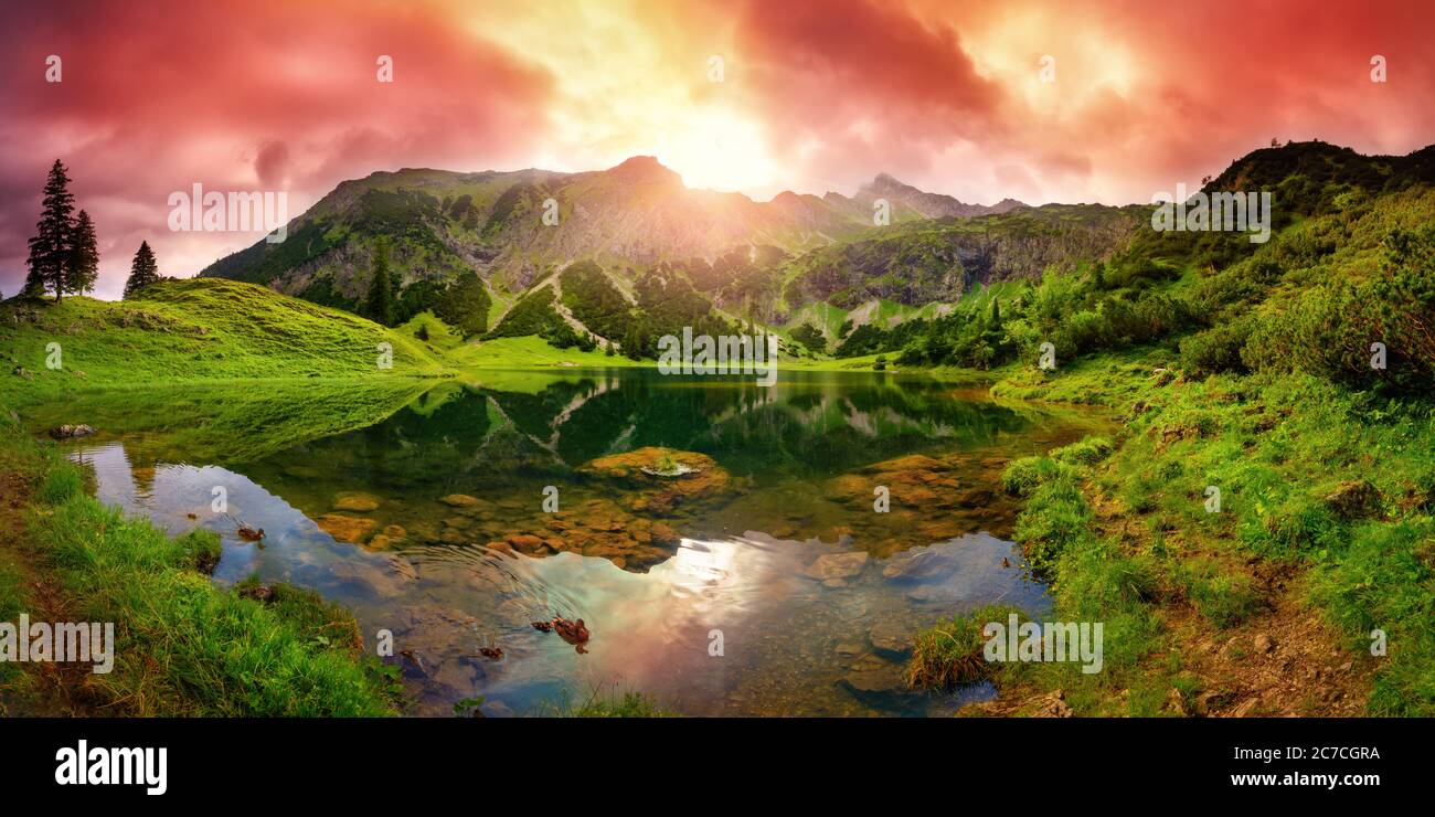 Alba spettacolare in un lago delle Alpi con montagne, nuvole rosse riflesse nelle acque cristalline e sentieri che conducono attraverso l'erba verde vibrante Foto Stock