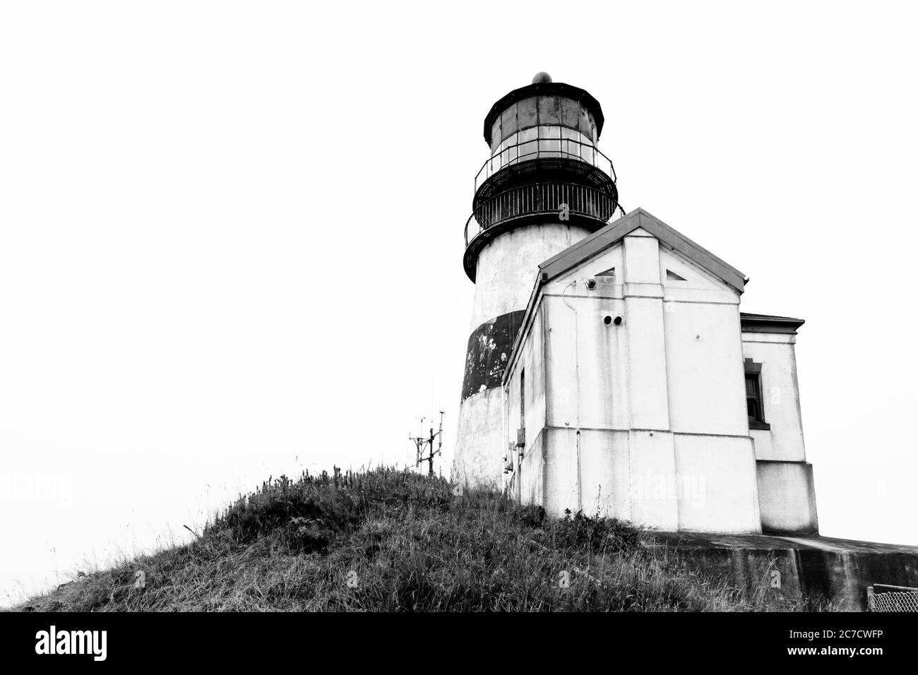 Scala di grigi a basso angolo di un faro vicino a un piccolo cabina su una scogliera Foto Stock