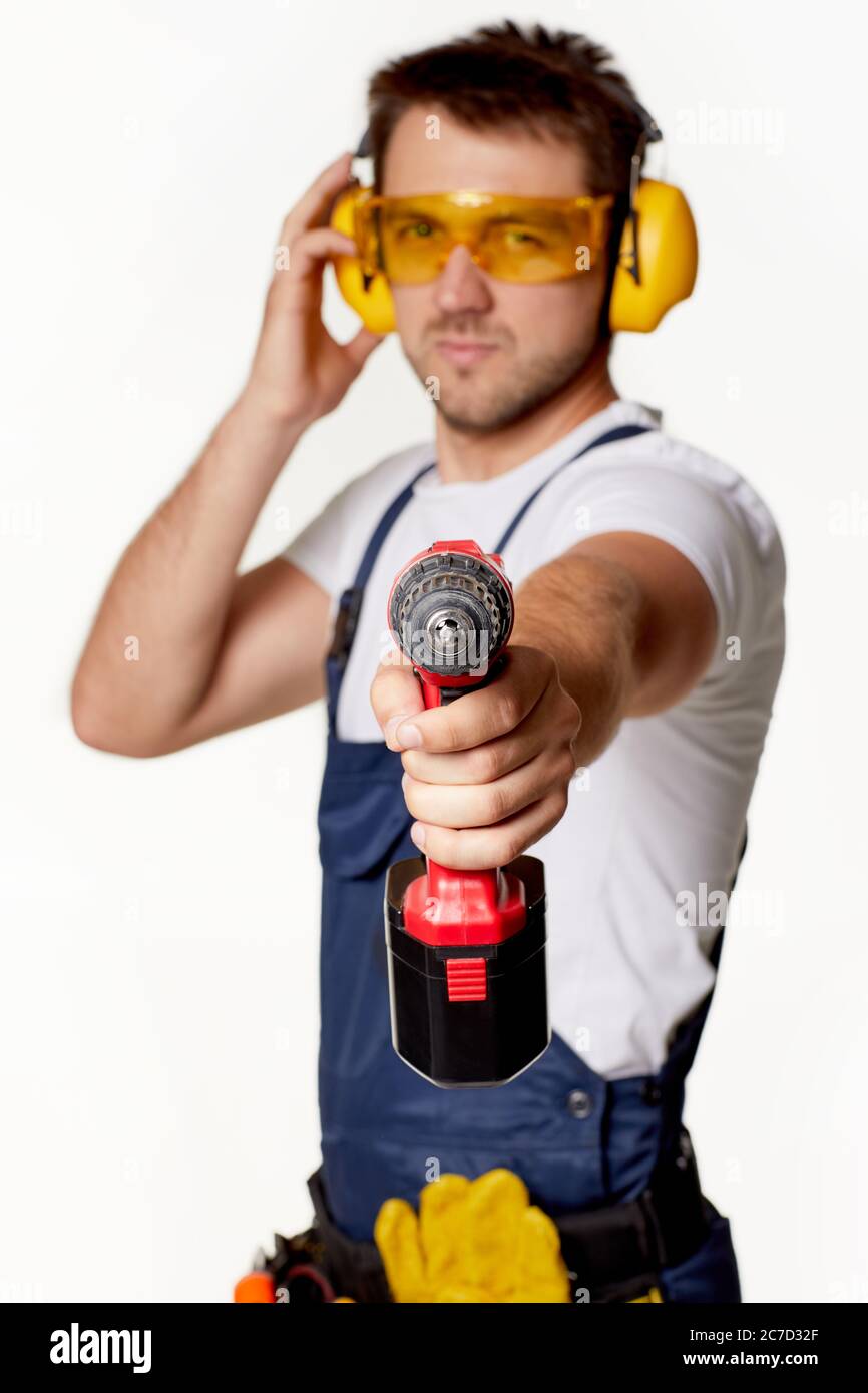 addetto alla riparazione caucasica in uniforme, occhiali di sicurezza e cuffie che tengono isolato il cacciavite senza fili su sfondo bianco. attenzione al cacciavite senza fili Foto Stock