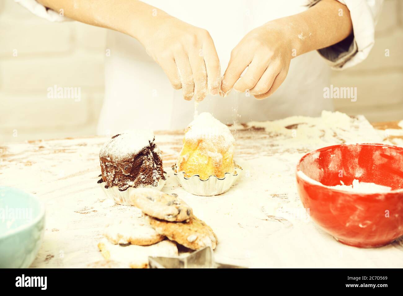 le mani maschili giovani del cuoco chef in uniforme bianca in piedi vicino al tavolo con ciotola rossa molta farina e cospargere gustose torte o muffin con zucchero in polvere, su fondo muro di mattoni Foto Stock