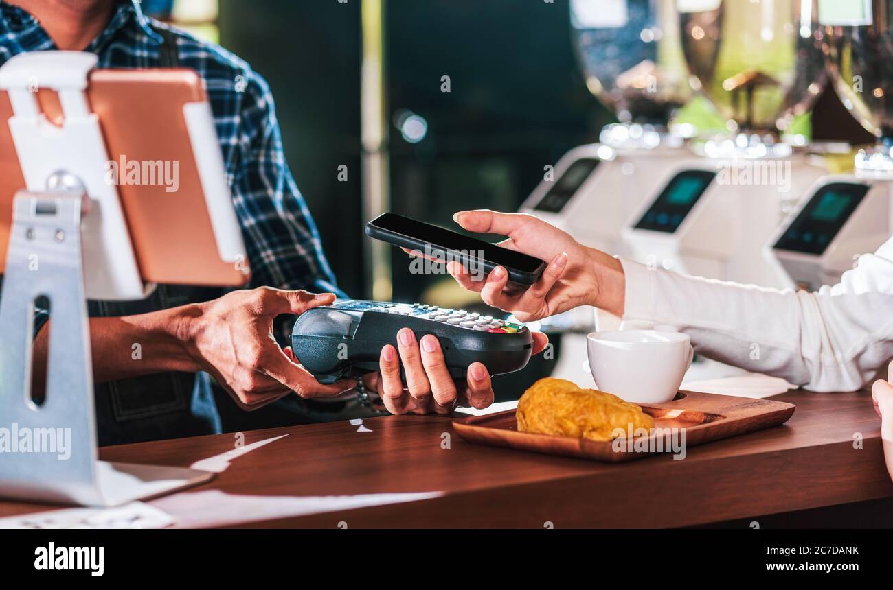 Closeup uomo asiatico mano di cliente che paga soldi via canale contactless da mobile banking applicazione a piccolo imprenditore al tavolo in negozio di caffè, Smal Foto Stock