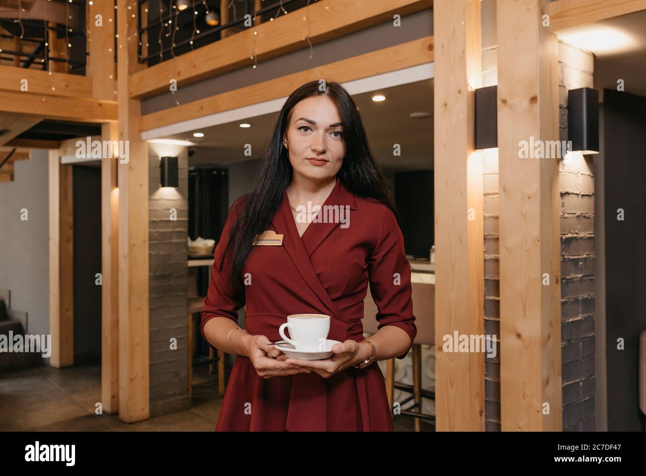 Una manager di un ristorante femminile sta tenendo una tazza di caffè in un ristorante. Un gentile proprietario di un caffè in un abito rubino sta aspettando un cliente. Foto Stock