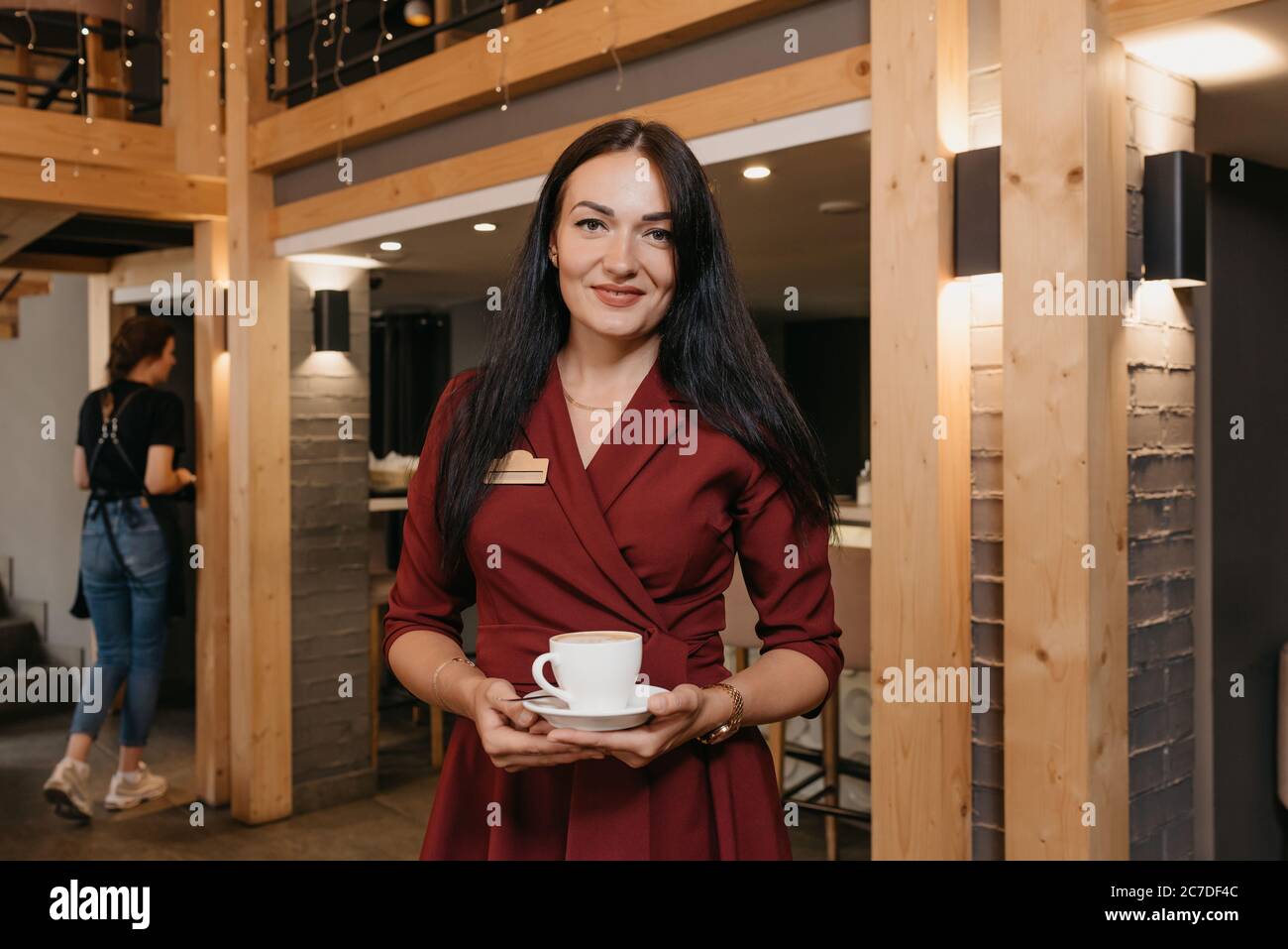 Una splendida manager di un ristorante femminile sta tenendo una tazza di caffè in un ristorante. Un gentile proprietario di un caffè in un abito rubino sta aspettando un cliente. Foto Stock