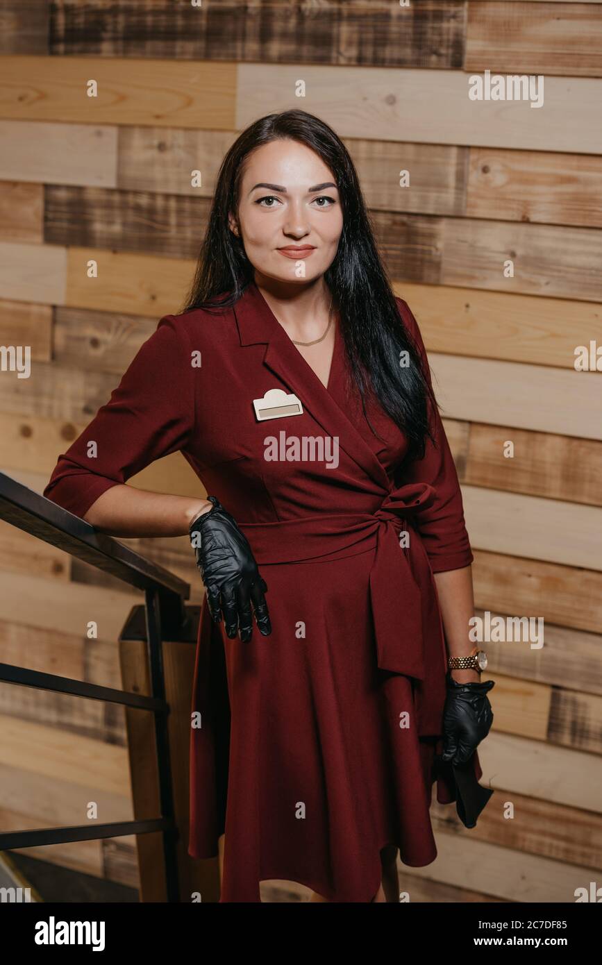 Una manager di ristorante femminile in guanti neri monouso che sorride e posa in una caffetteria. Un gentile proprietario di caffè in un abito rubino sta aspettando cl Foto Stock