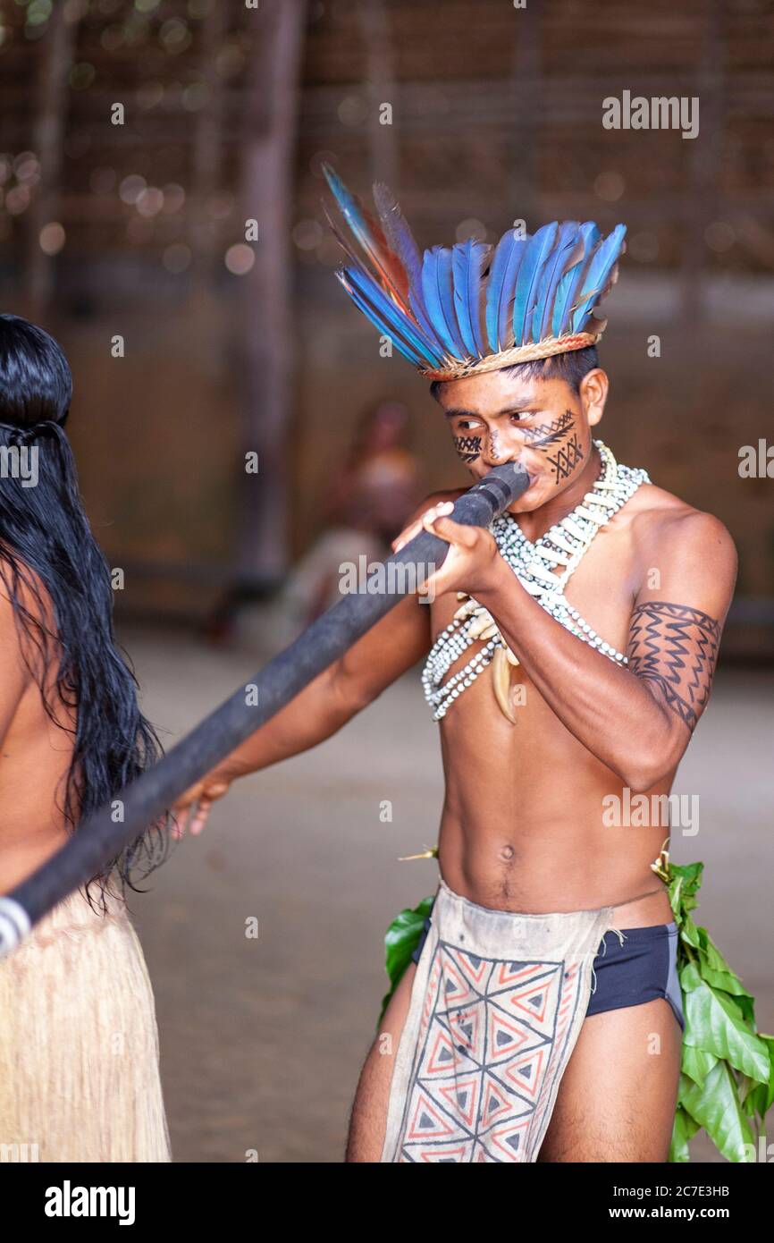 Un uomo indigeno amazzonico che indossa un copricapo piumato e una vernice per il viso suona uno strumento cerimoniale, celebrando le tradizioni culturali del Brasile Foto Stock