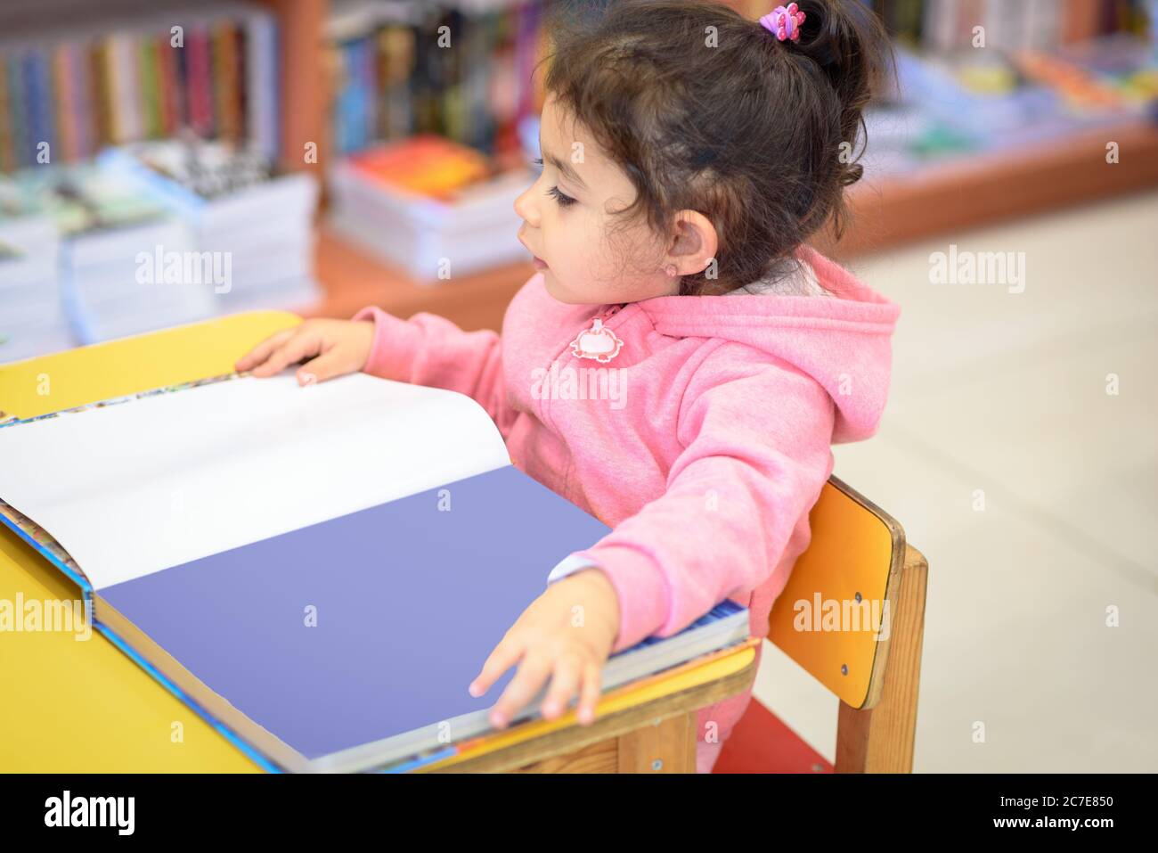 Bambina interni nella parte anteriore dei libri. Carino giovane bambino seduto su una sedia vicino al tavolo e la lettura del libro. Bambino legge in una libreria, circondato da colorati libri. Biblioteca, Shop, scaffalature in casa. Foto Stock