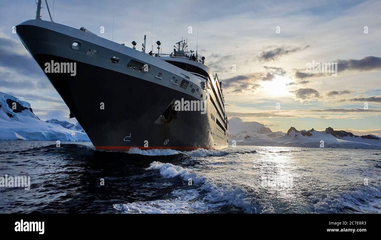 Nave da crociera nelle acque antartiche circondata da ghiacciai e montagne innevate con il sole serale sullo sfondo Foto Stock