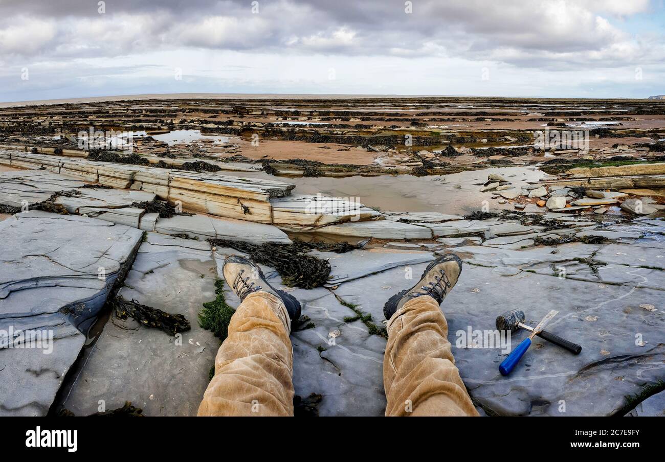 Gambe e stivali da uomo allungati con martello e scalpello messi a lato su una costa di ardesia britannica durante la bassa marea con fossili di ammonite nella roccia Foto Stock