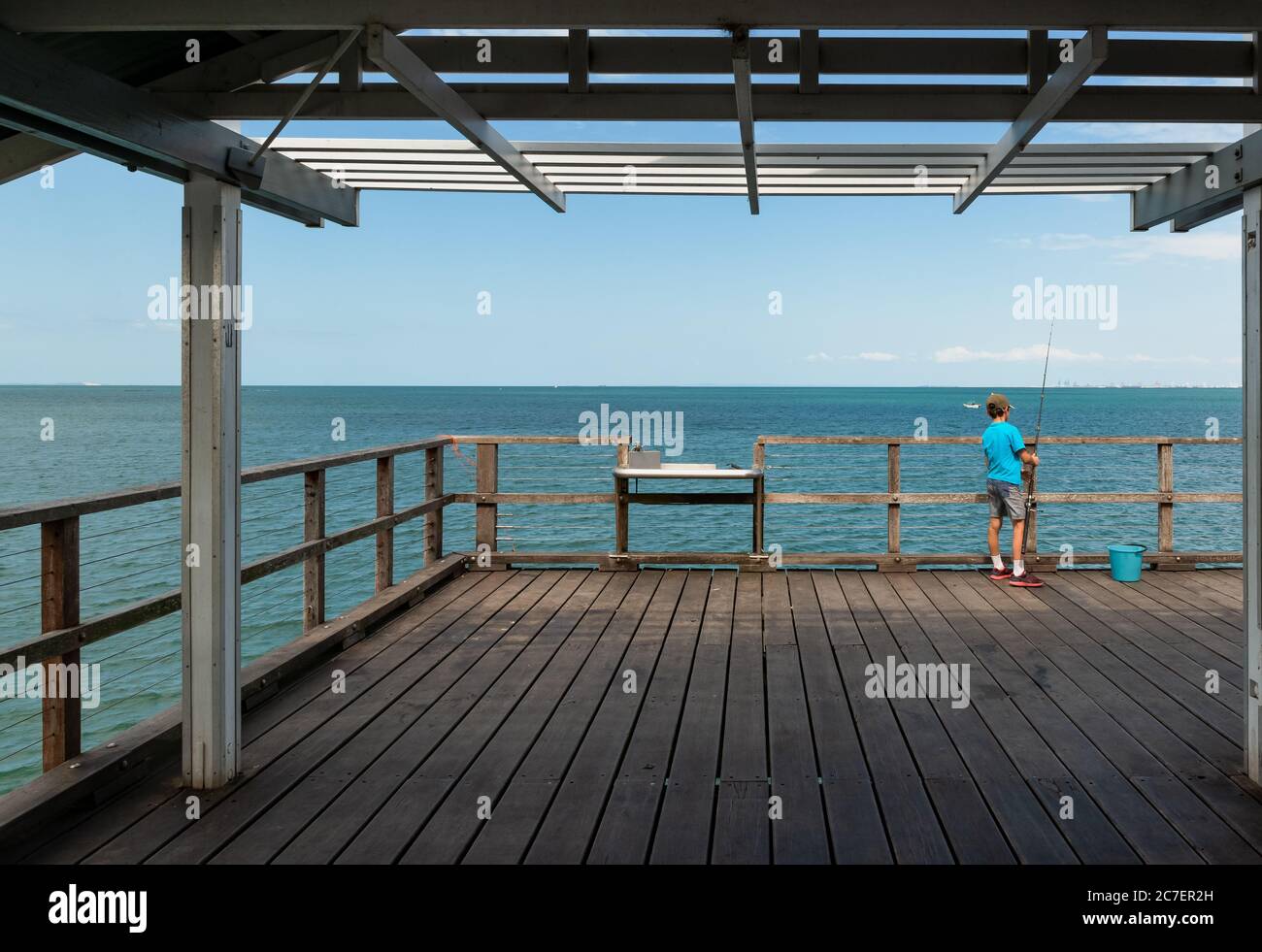 Ragazzo ottimisticamente sperando per un grande pesce in piedi un ponte di legno vicino a un secchio blu Foto Stock