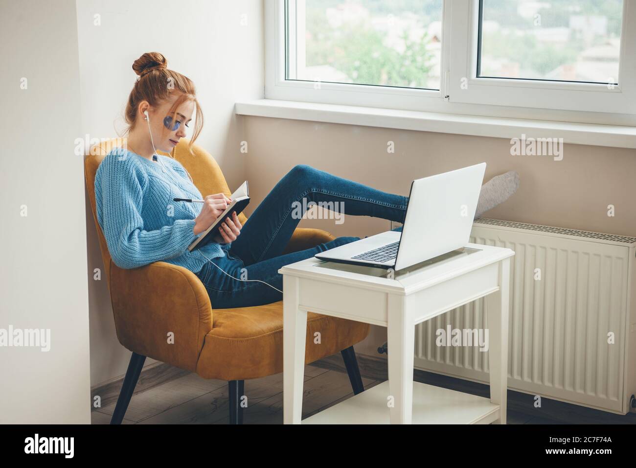 Donna caucasica con cocci e capelli rossi che indossano macchie di idrogel oculari mentre si scrive nel libro e si utilizza un computer portatile con auricolari Foto Stock