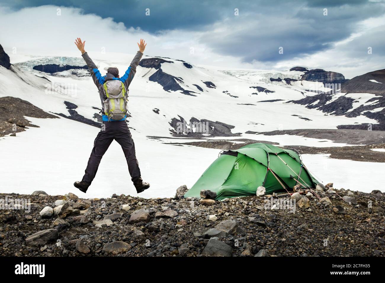 Uomo escursionista con zaino e tenda saltando con braccia allungate. Campeggio in ghiacciai di montagna neve e vento tempesta. Thorisdalur, Thorisjokull, Islanda Foto Stock