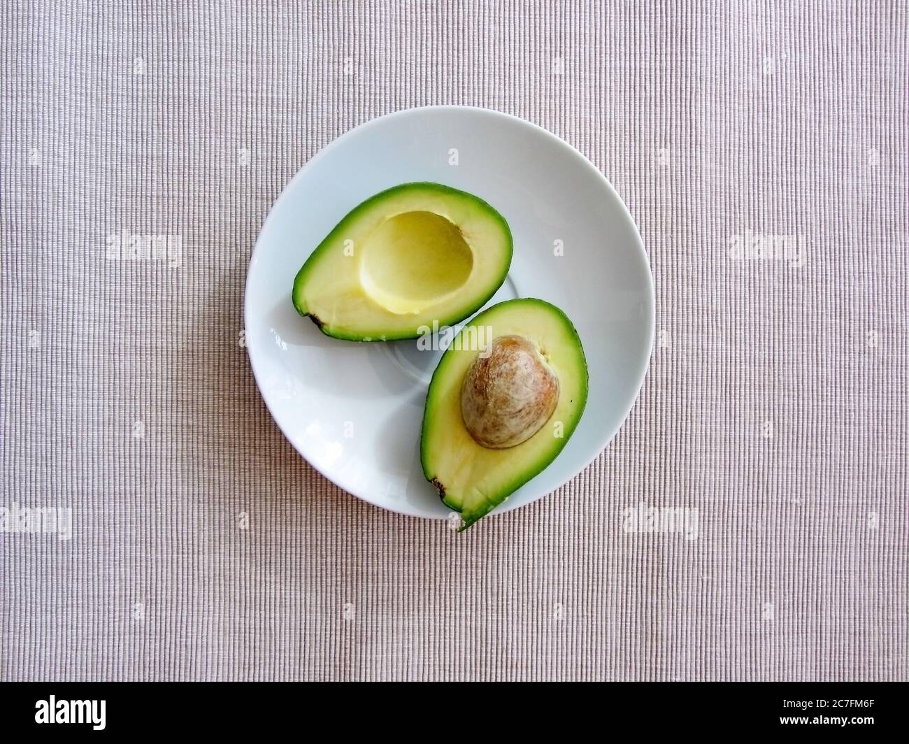 Le metà fresche di avocado biologico, una con osso, giacciono su un piatto bianco su una tela a righe di cotone grigio, giacitura piatta. Concetto di cibo sano.. Foto Stock