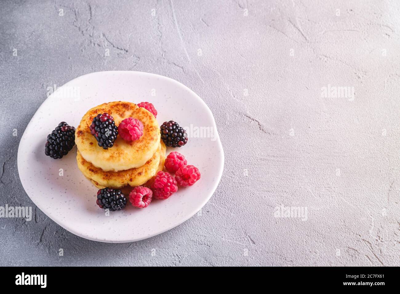 Frittelle al formaggio di cottage, frittelle di cagliata dessert con lampone e frutti di mora in piastra su fondo di pietra in cemento, angolo di vista spazio copia Foto Stock