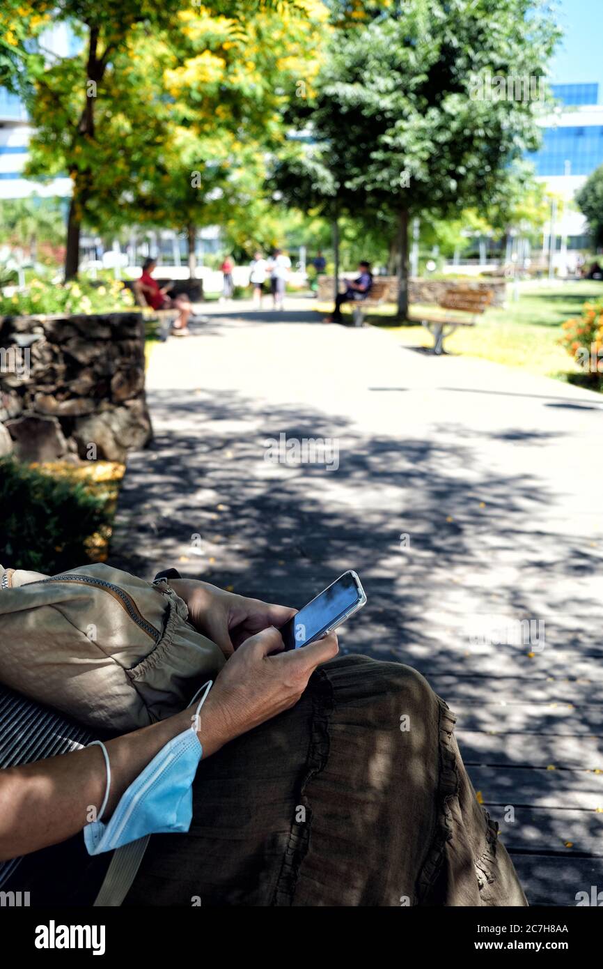 Vista ravvicinata delle mani di una donna matura con smartphone e maschera seduta su una panchina in un parco Foto Stock